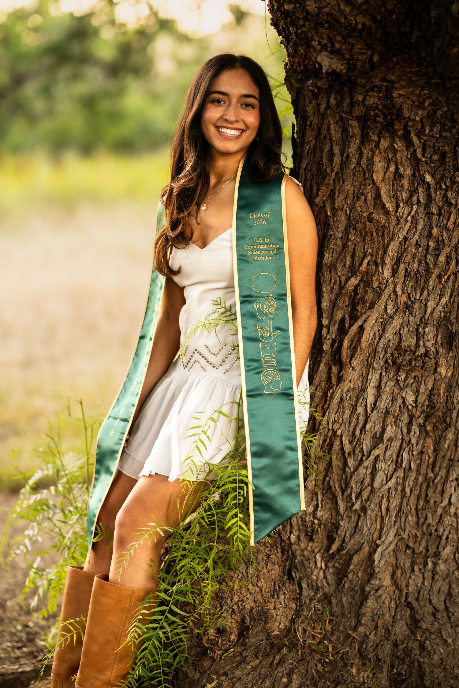 Smiling graduate in white dress with green sash leaning against oak tree in Brentwood, CA.