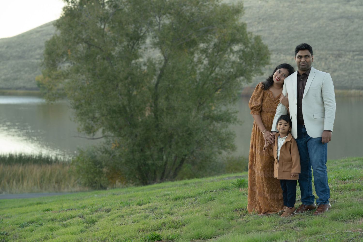 Family of three posing outdoors near scenic lake and green hills during golden hour portrait session.