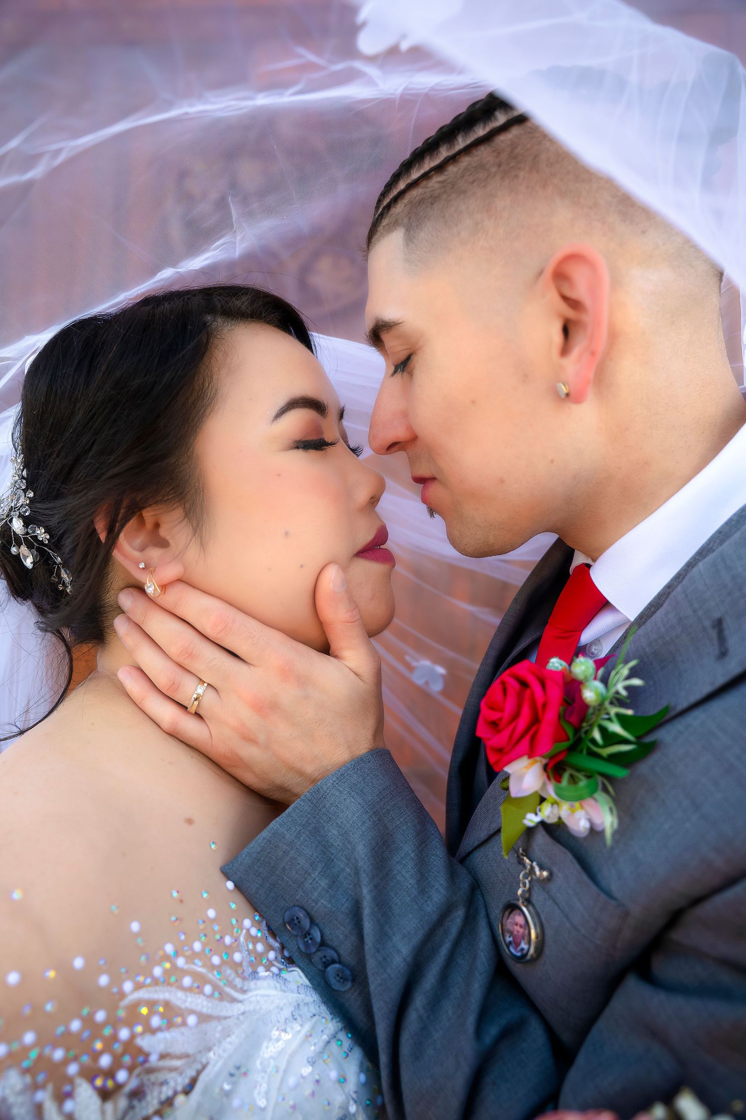 Bride and groom sharing intimate moment under veil with red rose boutonniere.