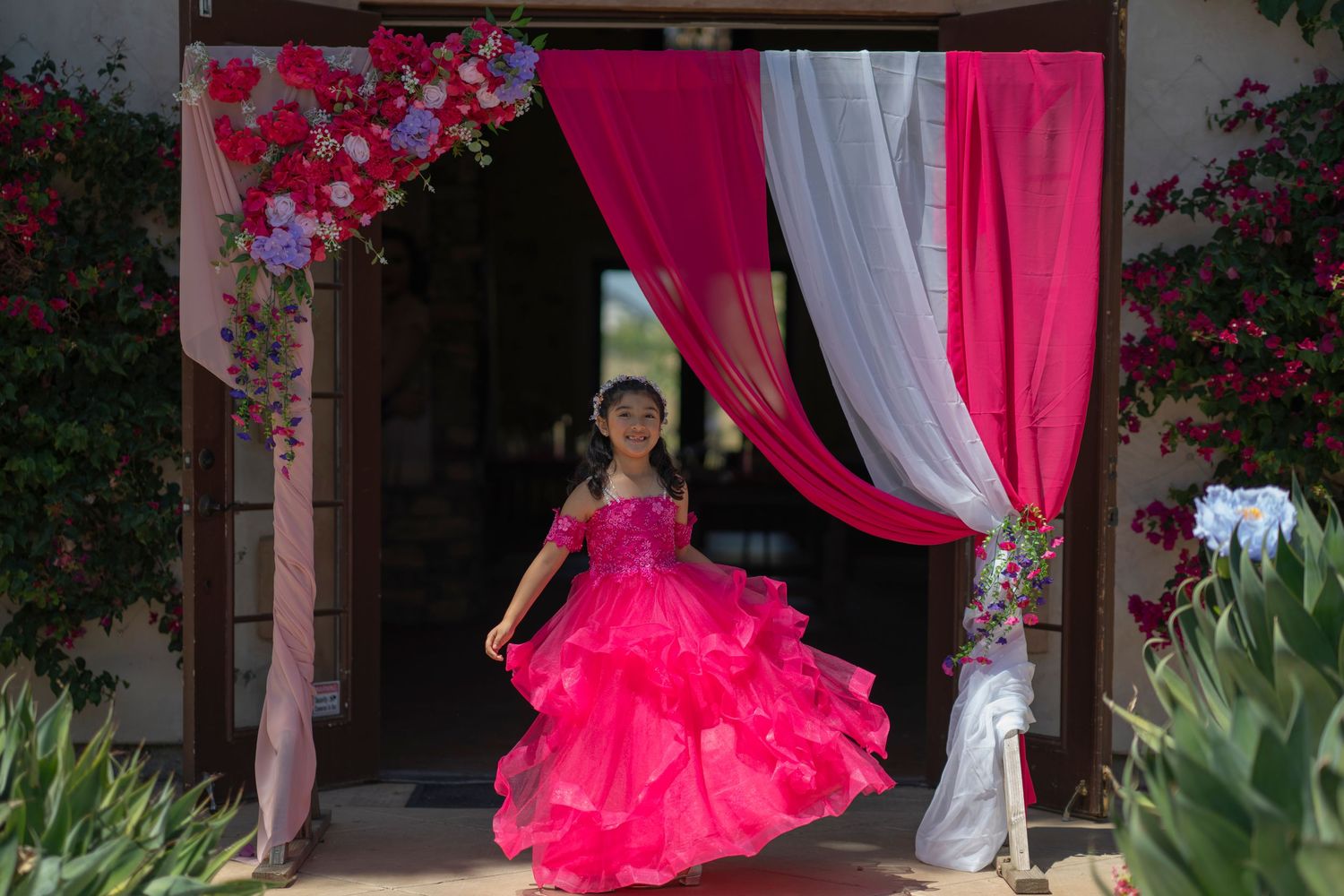 Young girl in a vibrant pink ruffled ball gown twirling under a floral arch with pink and white draping.