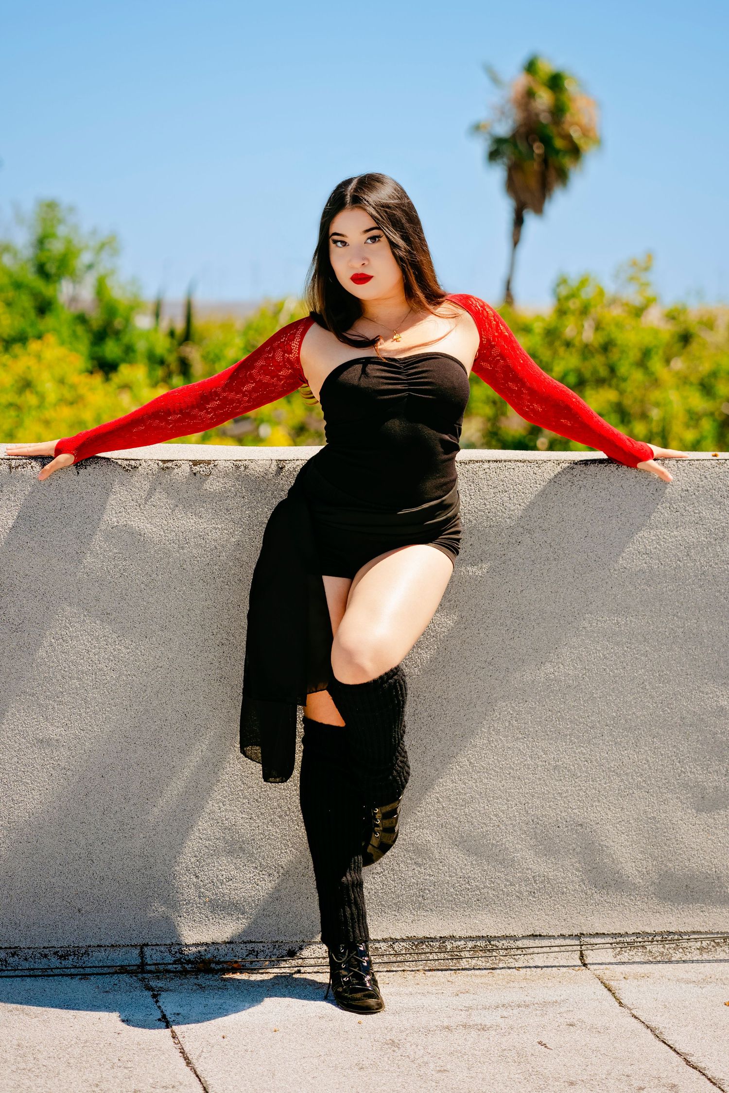 Woman in black dress and red lace sleeves posing against a concrete wall outdoors on a sunny day.