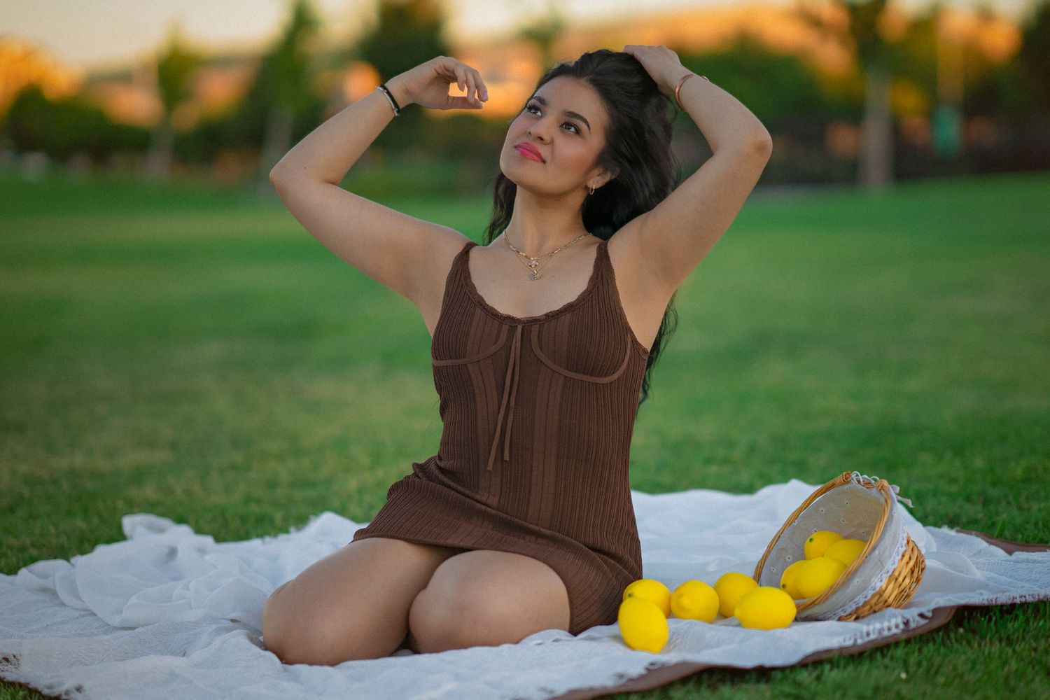 Woman in brown dress sitting on blanket in park with yellow flowers and woven bag at golden hour.