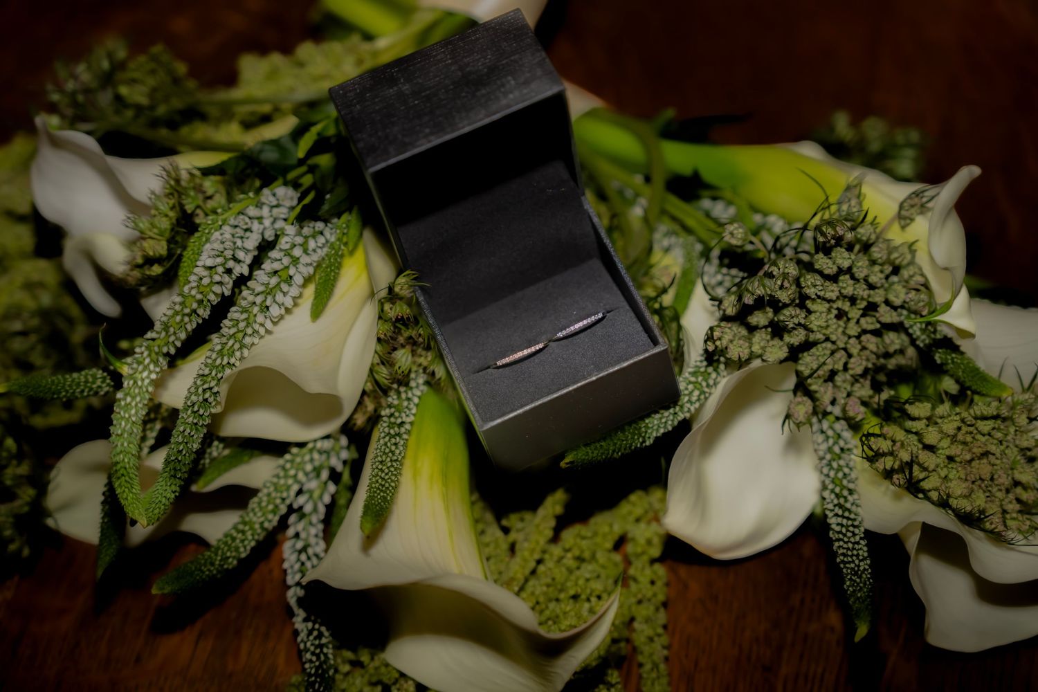 Wedding ring in black box displayed on white calla lily and green floral bouquet arrangement.