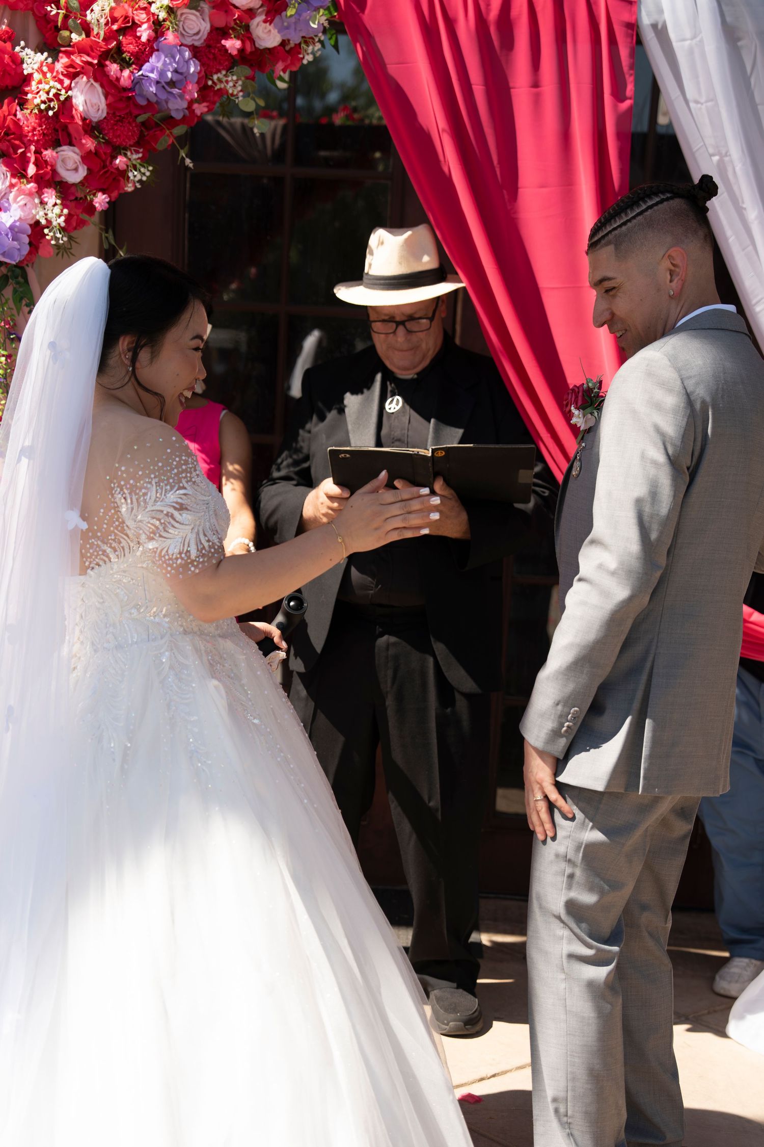 Bride placing ring on groom's finger during outdoor wedding ceremony with officiant and floral arch.