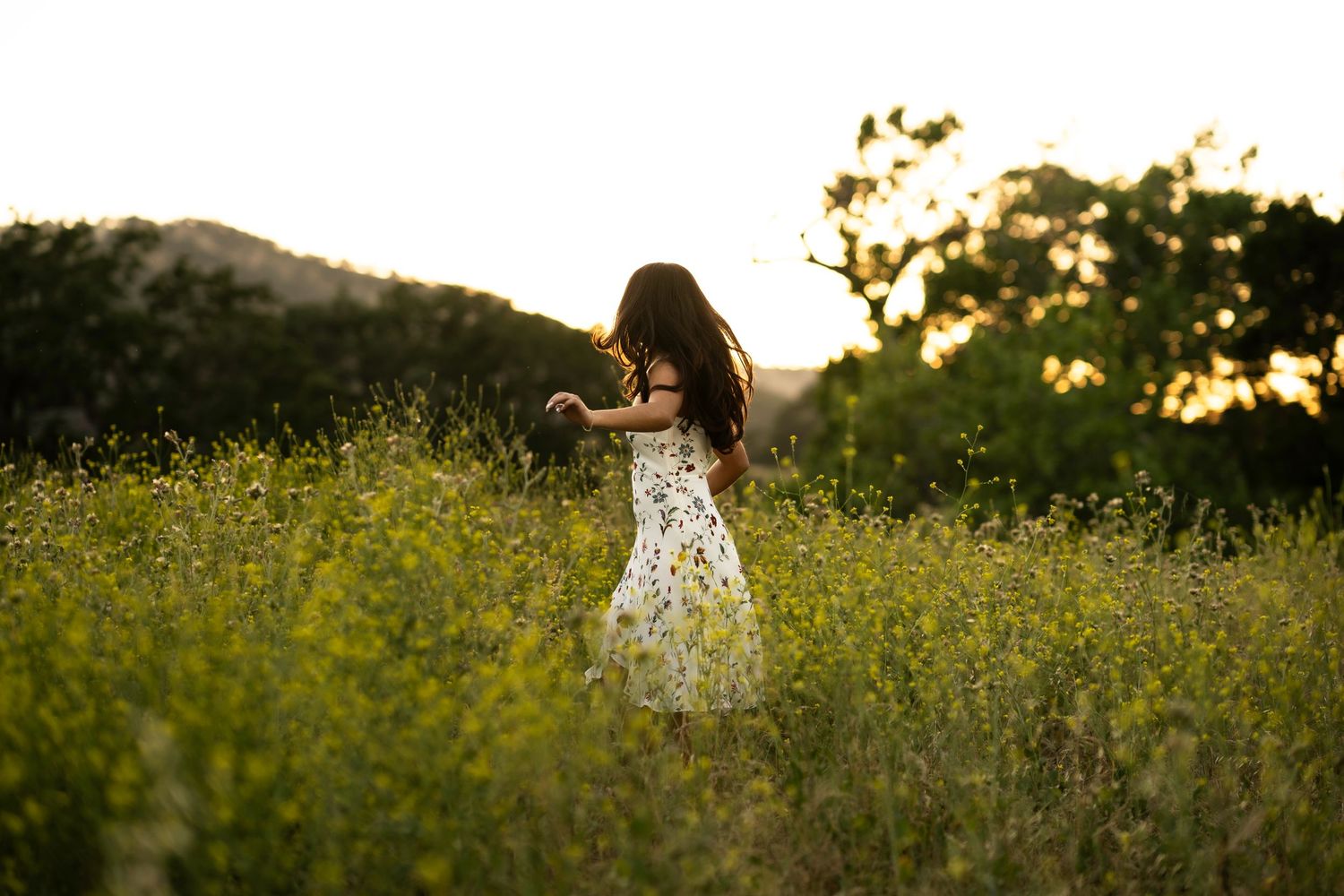 Young girl in floral dress walking through golden wildflower meadow at sunset in Brentwood, CA..