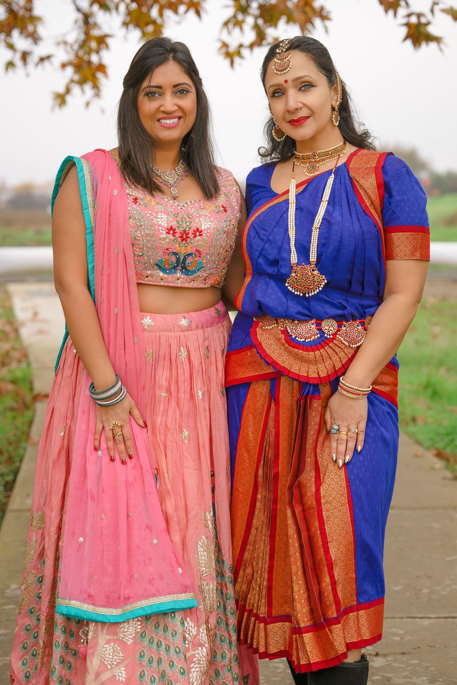Two women in traditional Indian attire, one in pink lehenga and one in blue saree, posing outdoors.