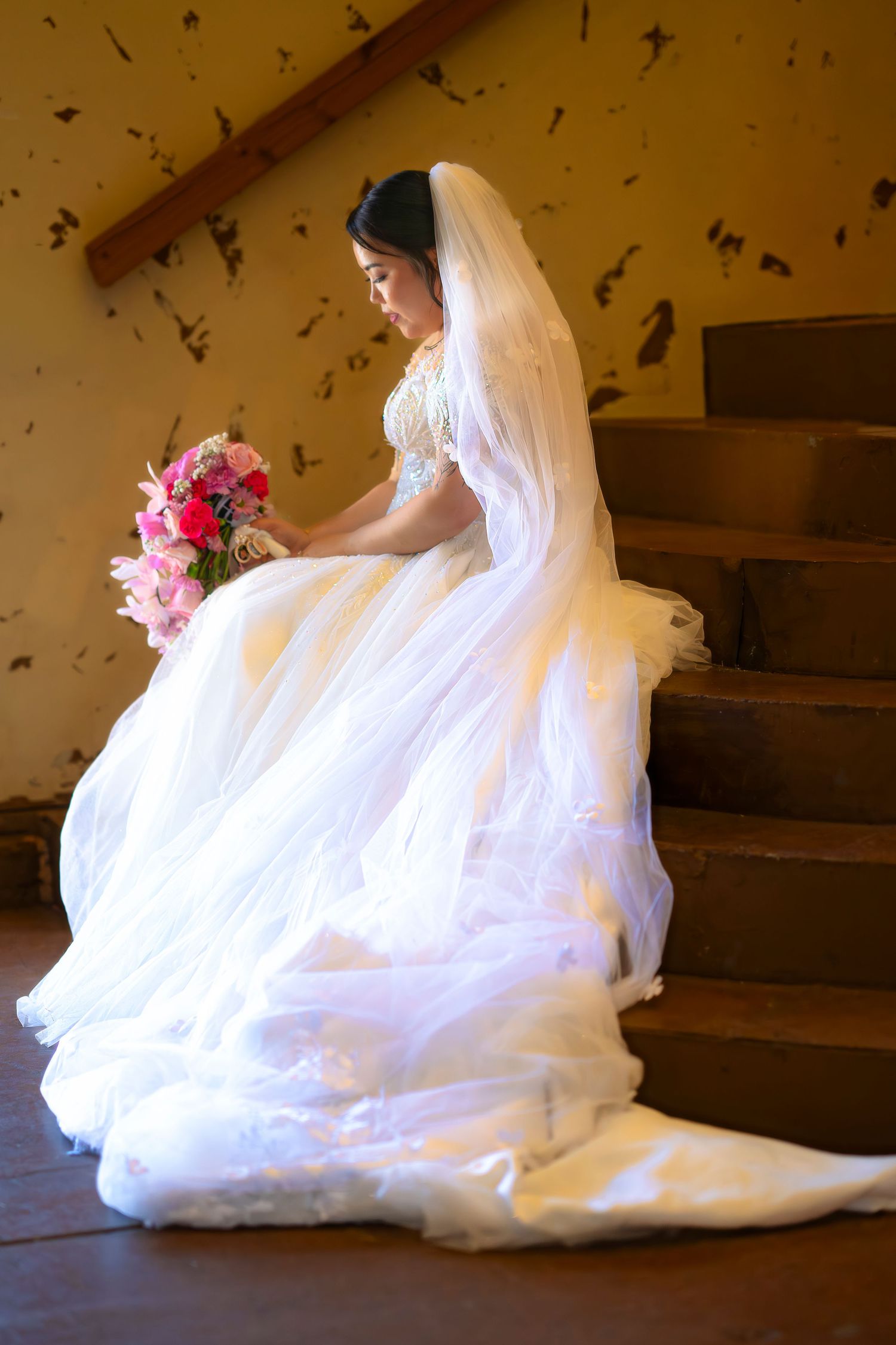 Bride in white ball gown with veil sitting on stairs holding pink bouquet.