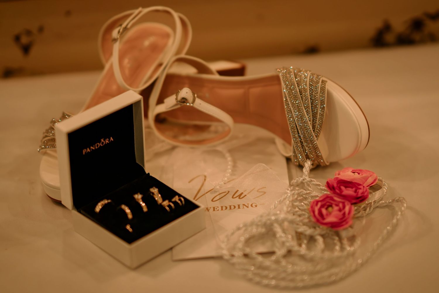 Elegant wedding flat lay featuring pink heels, Pandora jewelry box with gold rings, pearl necklace, and pink roses on white background.
