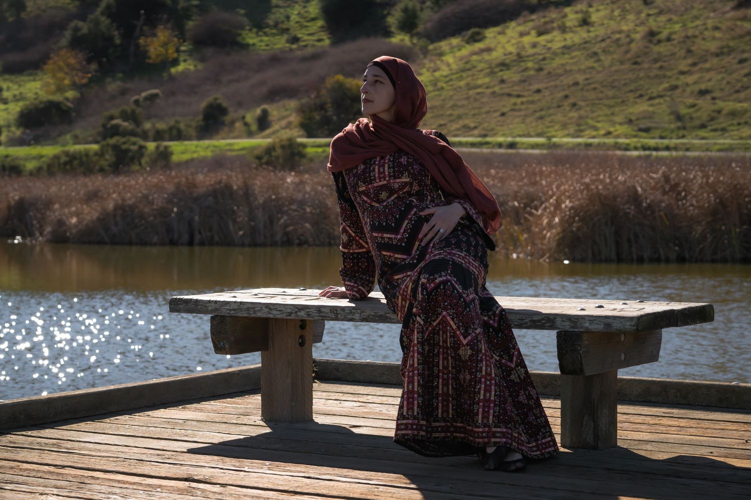 Woman in red hijab and patterned maxi dress sitting on a dock bench by a serene lake. Maternity Photo.