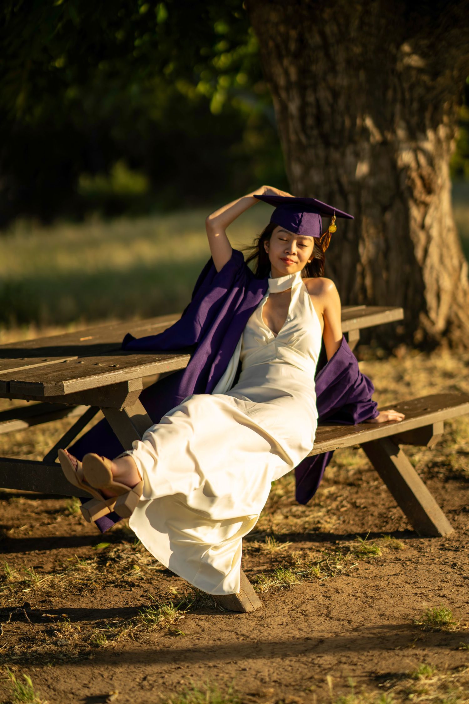 Woman in purple graduation cap and gown relaxing on a picnic bench outdoors in warm sunlight.