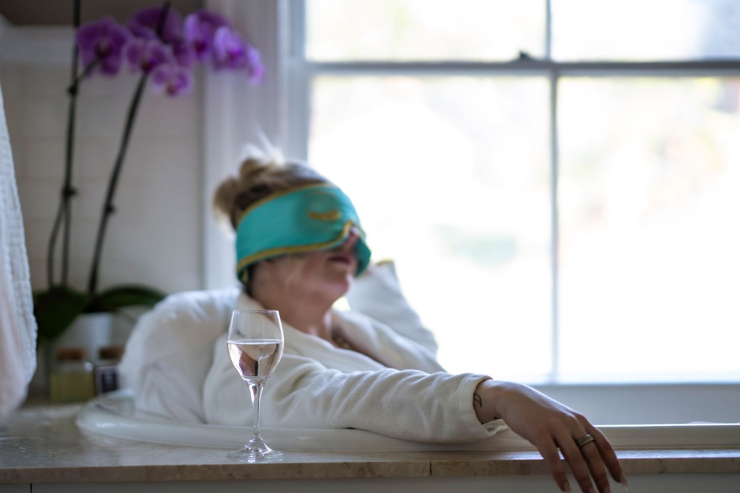 Relaxed woman wearing teal sleep mask beside wine glass near orchid window. Entrepreneur Life.