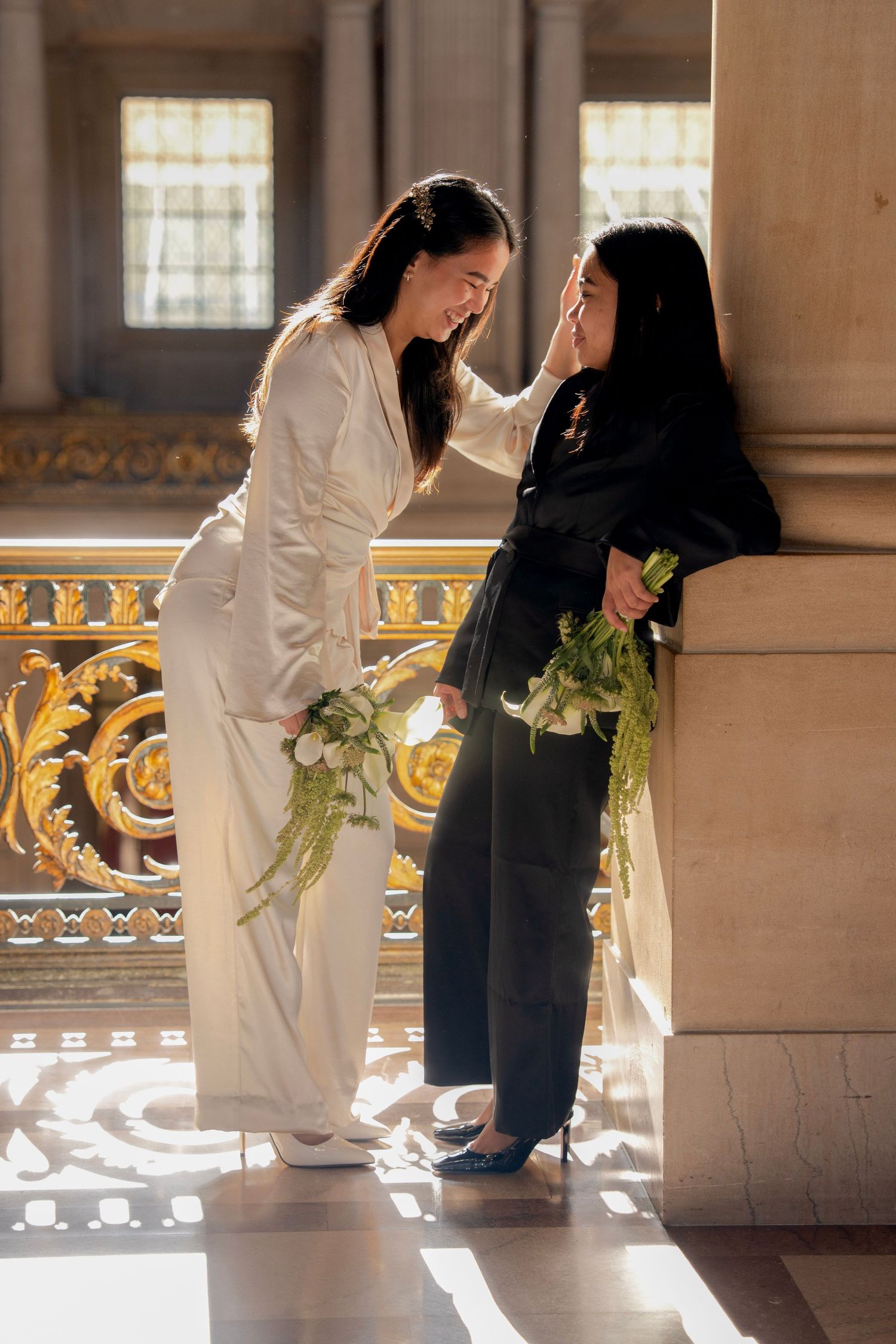 Two brides in white and black outfits hold floral bouquets while sharing a tender moment in an ornate golden hall in San Francisco City Hall