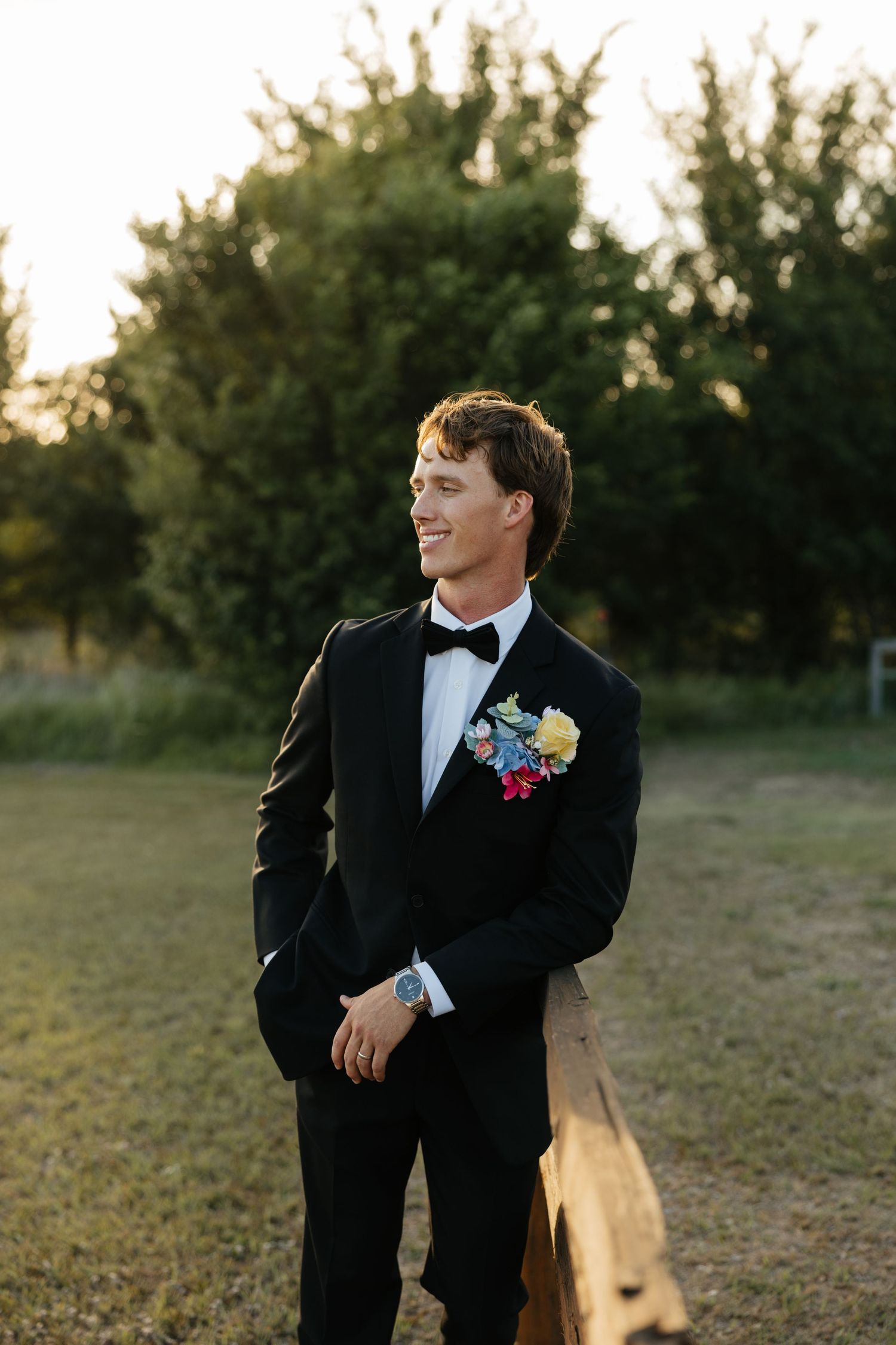 Dapper groom in a black tuxedo with colorful boutonniere smiles outdoors during golden hour at his wedding.