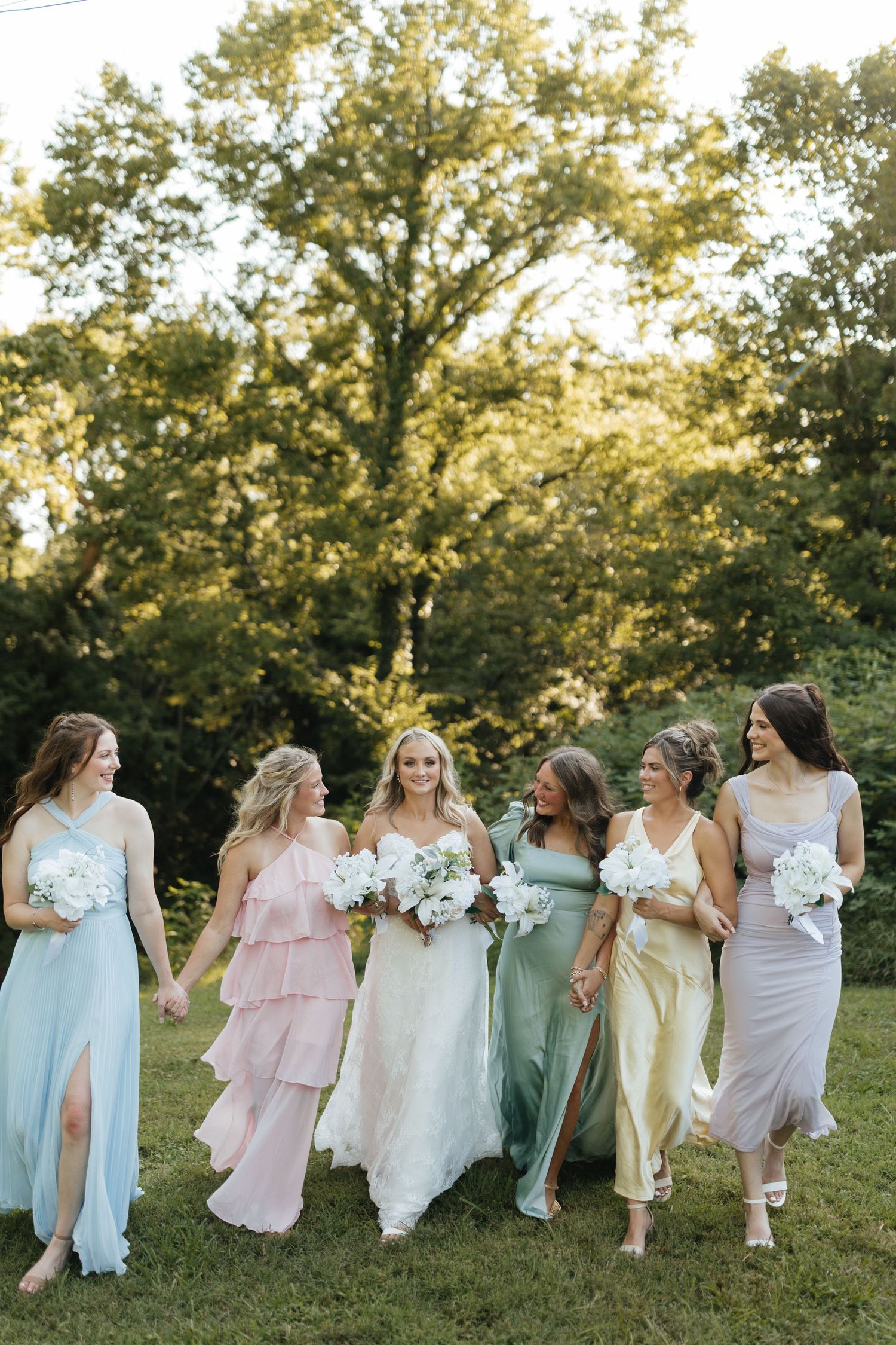 Bride and five bridesmaids in pastel mismatched gowns walking outdoors holding white bouquets.