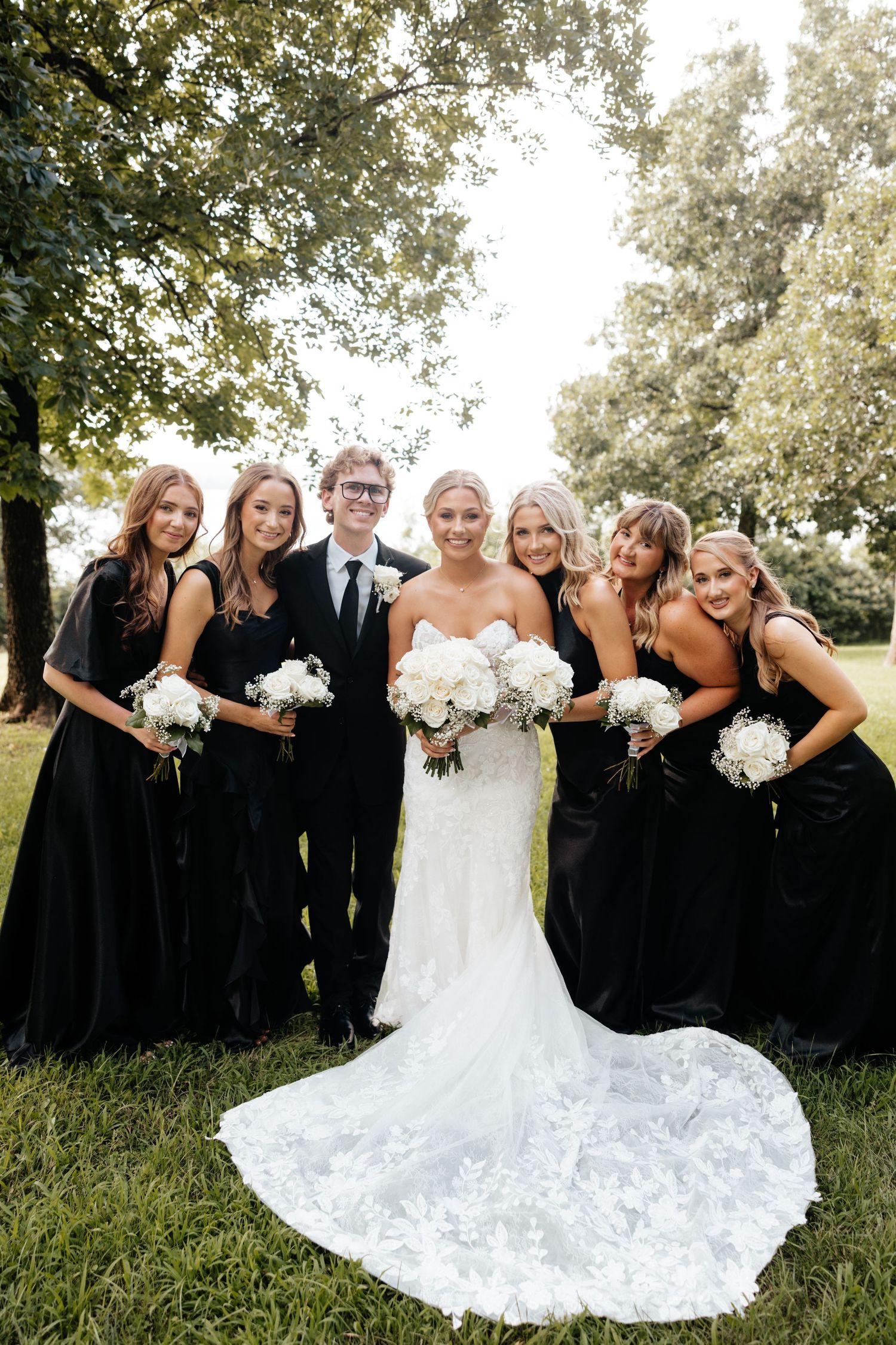Bride in lace wedding gown with bridesmaids in black dresses and groomsman holding white bouquets outdoors.
