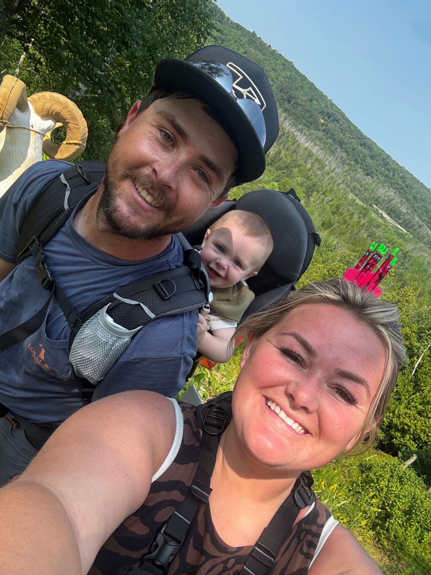 Family selfie with man carrying baby in backpack carrier and smiling woman outdoors.