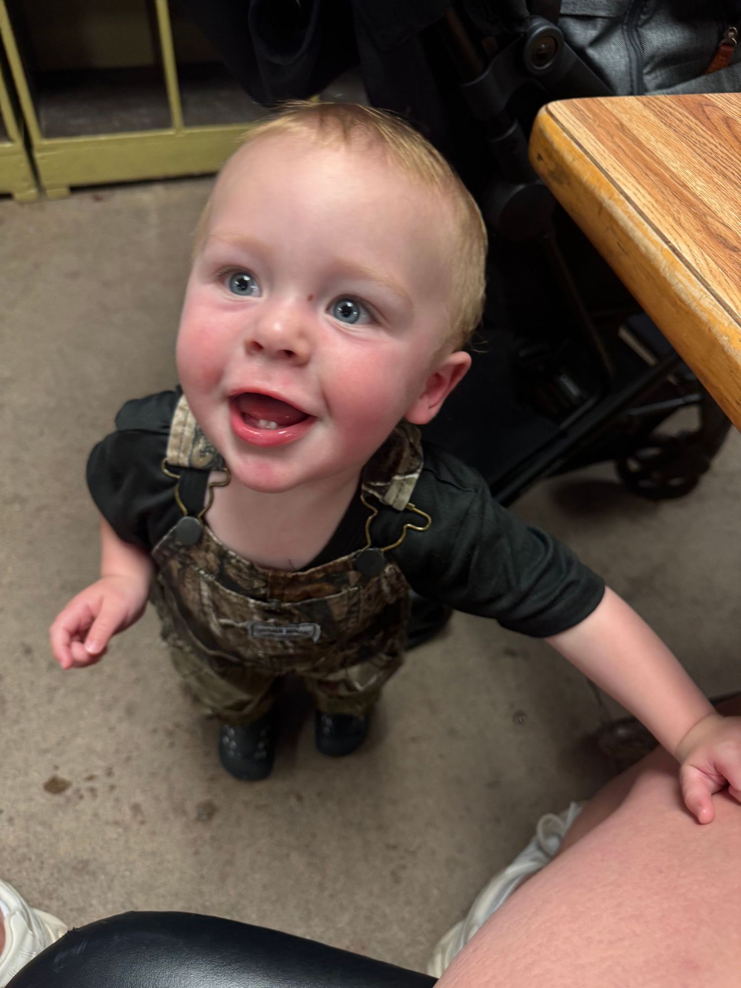 Happy toddler in camo overalls smiling up at camera indoors.