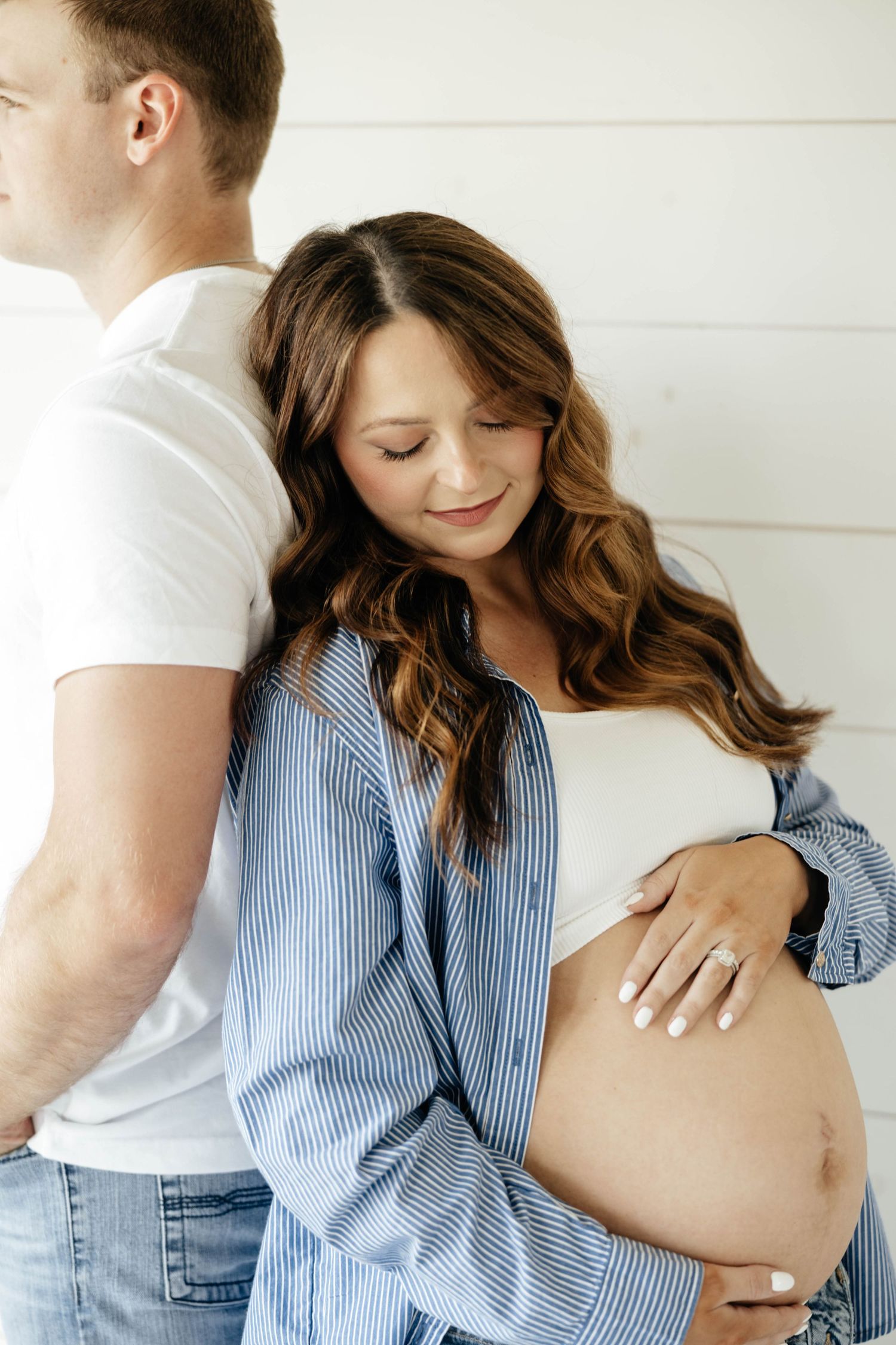 Pregnant woman in striped shirt tenderly holding baby bump while leaning against partner indoors.
