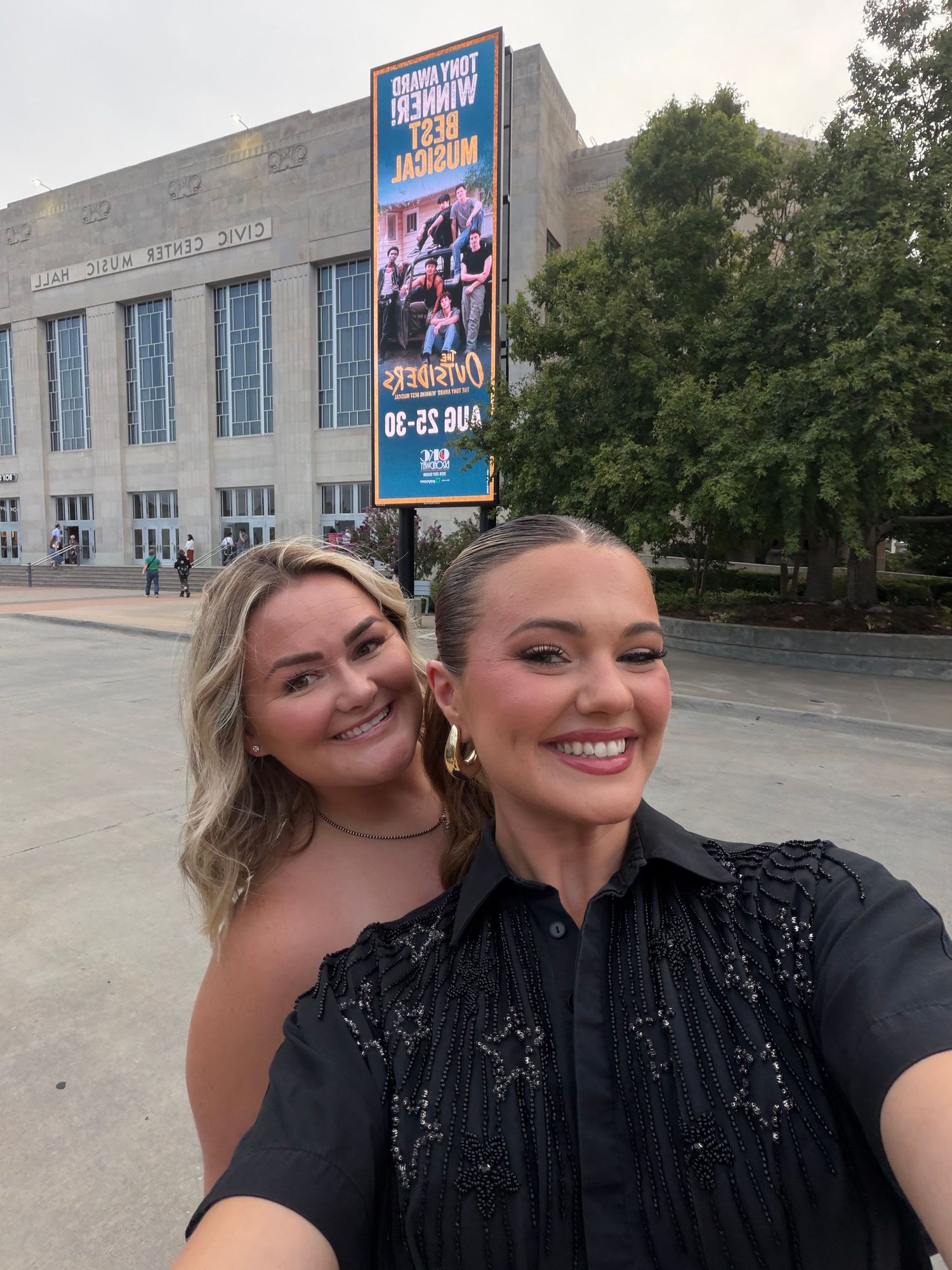 Two women smiling for a selfie outside a venue with a concert marquee sign behind them.