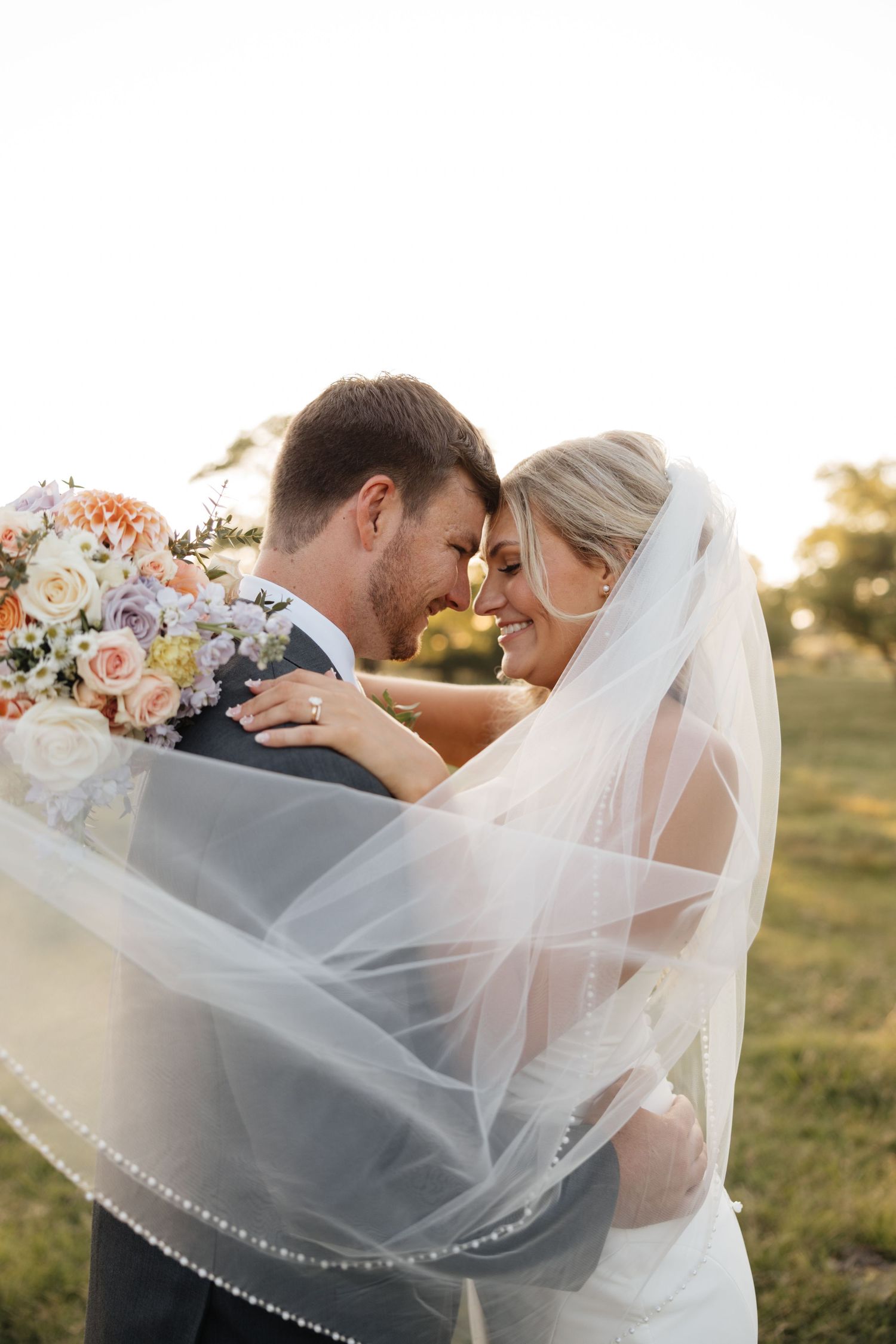 Bride and groom touching foreheads romantically under flowing veil at golden hour outdoor wedding.