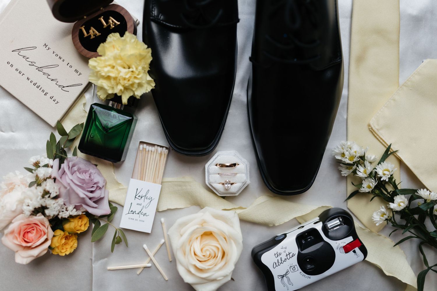 Groom wedding details flat lay with black dress shoes, cologne, roses, and disposable camera.