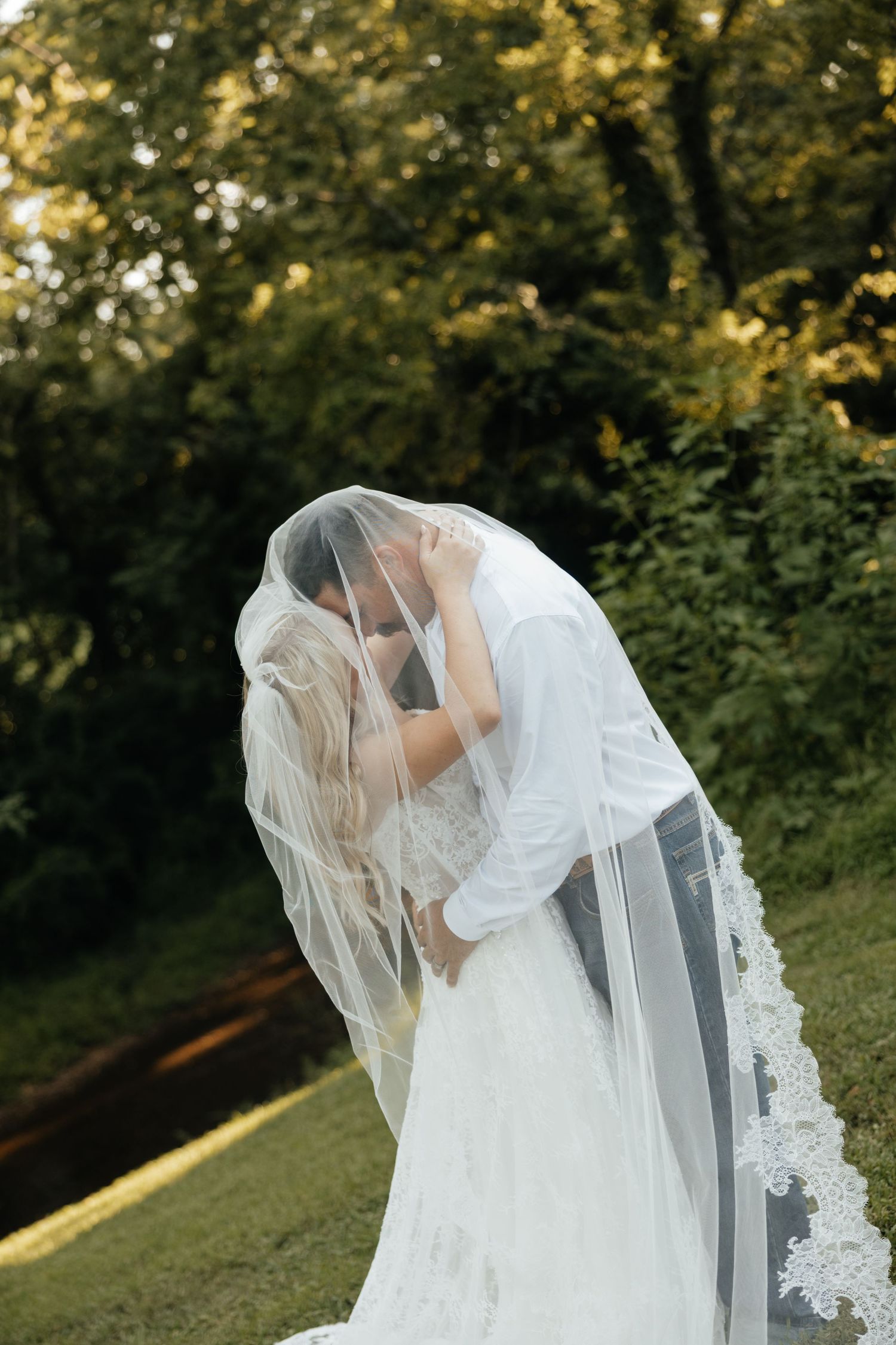 Bride and groom sharing a kiss hidden under flowing lace wedding veil surrounded by lush green trees.