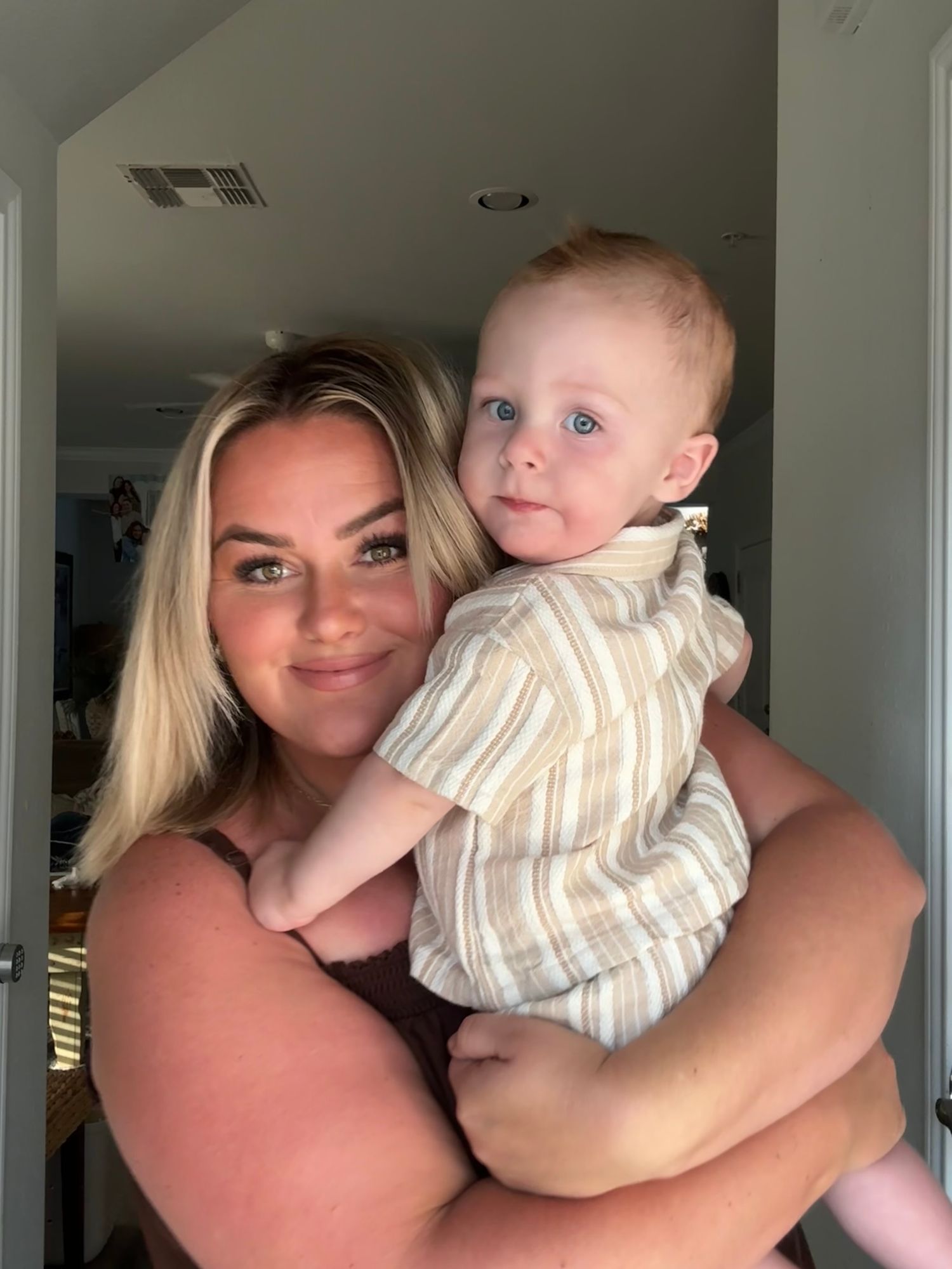 Smiling blonde woman holds blue-eyed baby boy in striped shirt indoors.