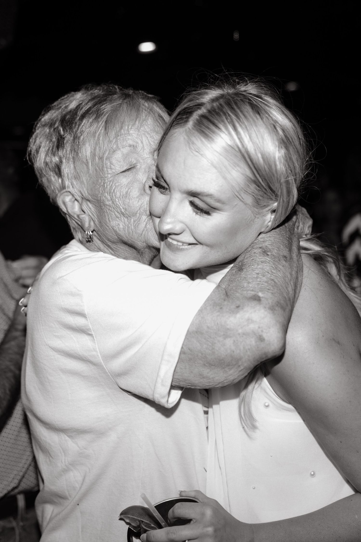 Black and white photo of elderly woman kissing blonde bride on cheek at wedding reception.