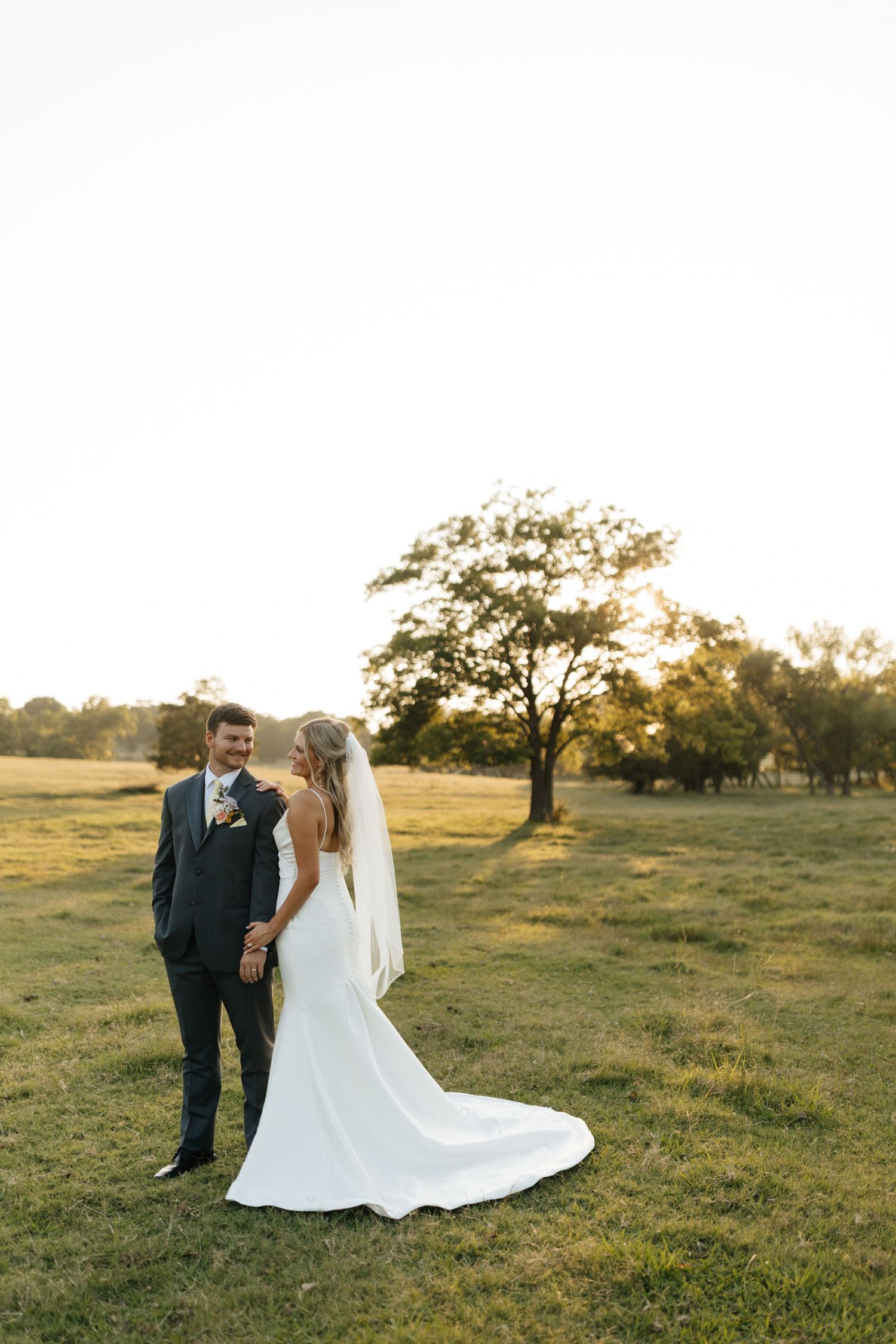 Bride in elegant white gown with cathedral veil stands with groom in gray suit during golden hour outdoor portraits.
