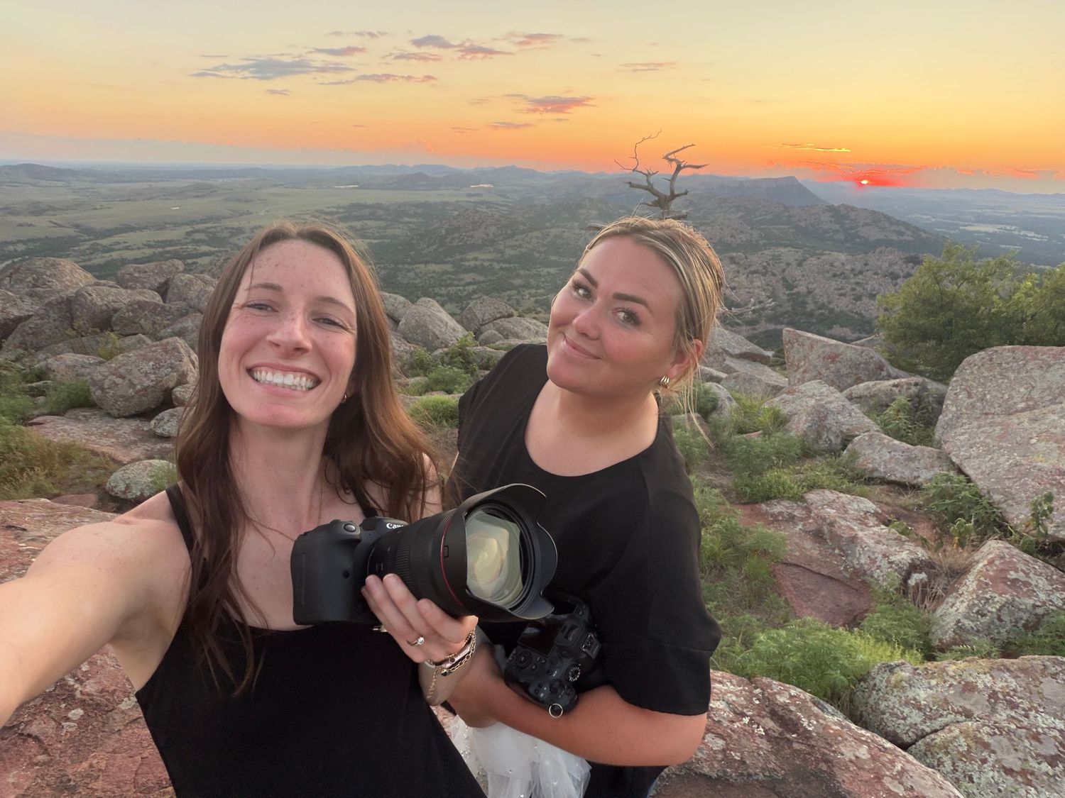 Two smiling women taking selfie with camera at rocky summit during vibrant sunset.