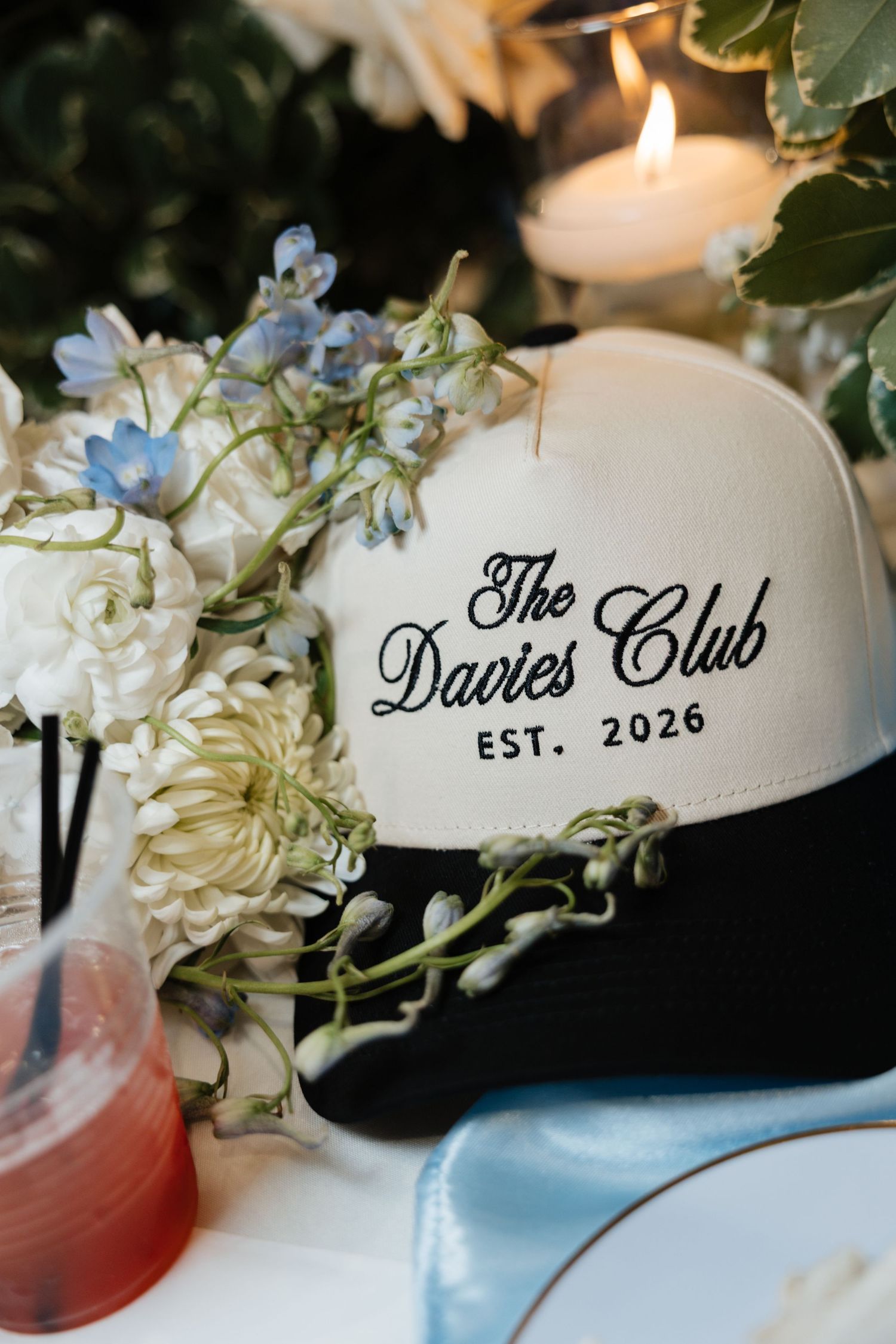 Custom trucker hat reading The Davies Club Est. 2026 displayed among white floral wedding centerpiece decorations.