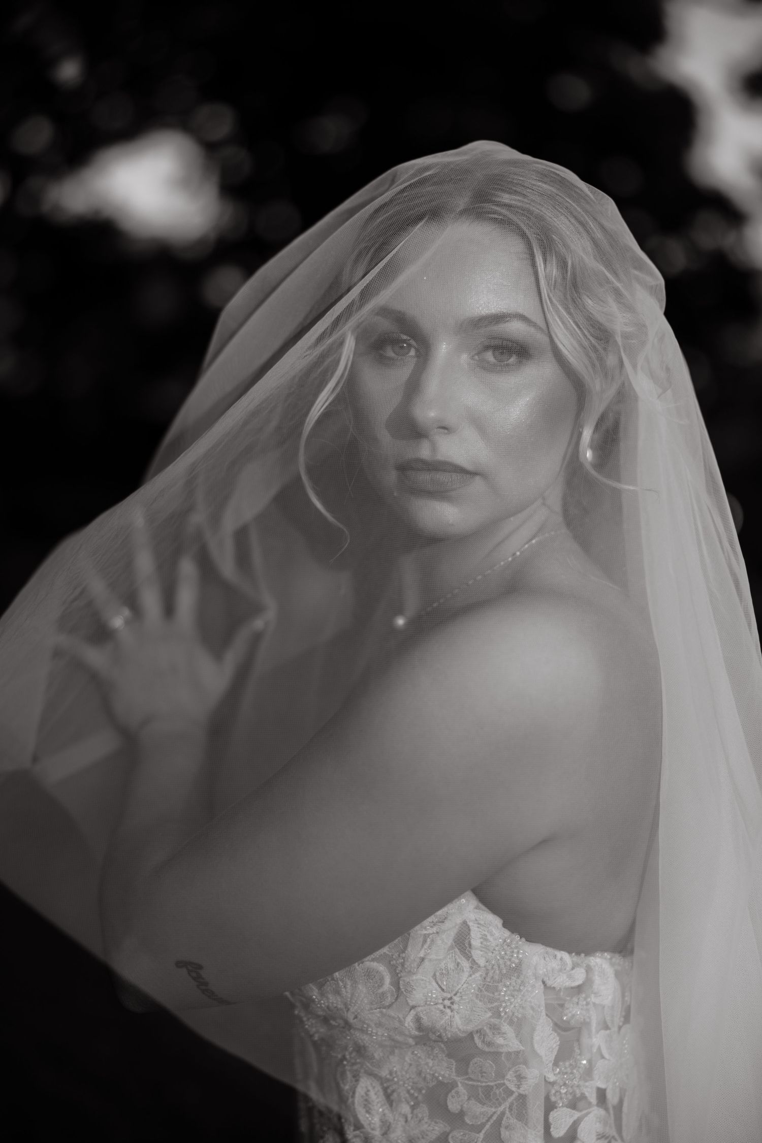 Elegant black and white bridal portrait of blonde bride wearing lace gown and flowing veil outdoors.