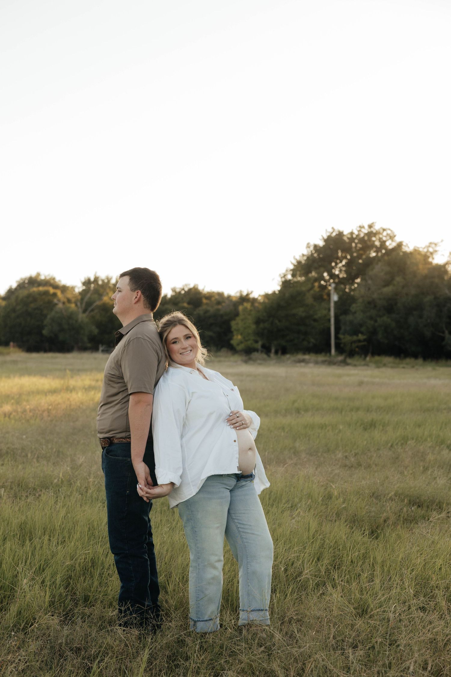 Pregnant woman and partner standing back to back in golden sunlit grassy field.