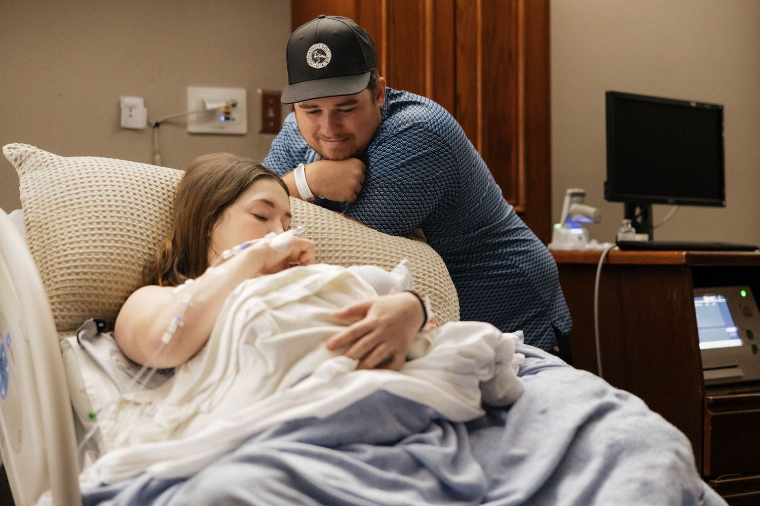 New father tenderly embraces exhausted mother holding newborn baby in hospital room after delivery.