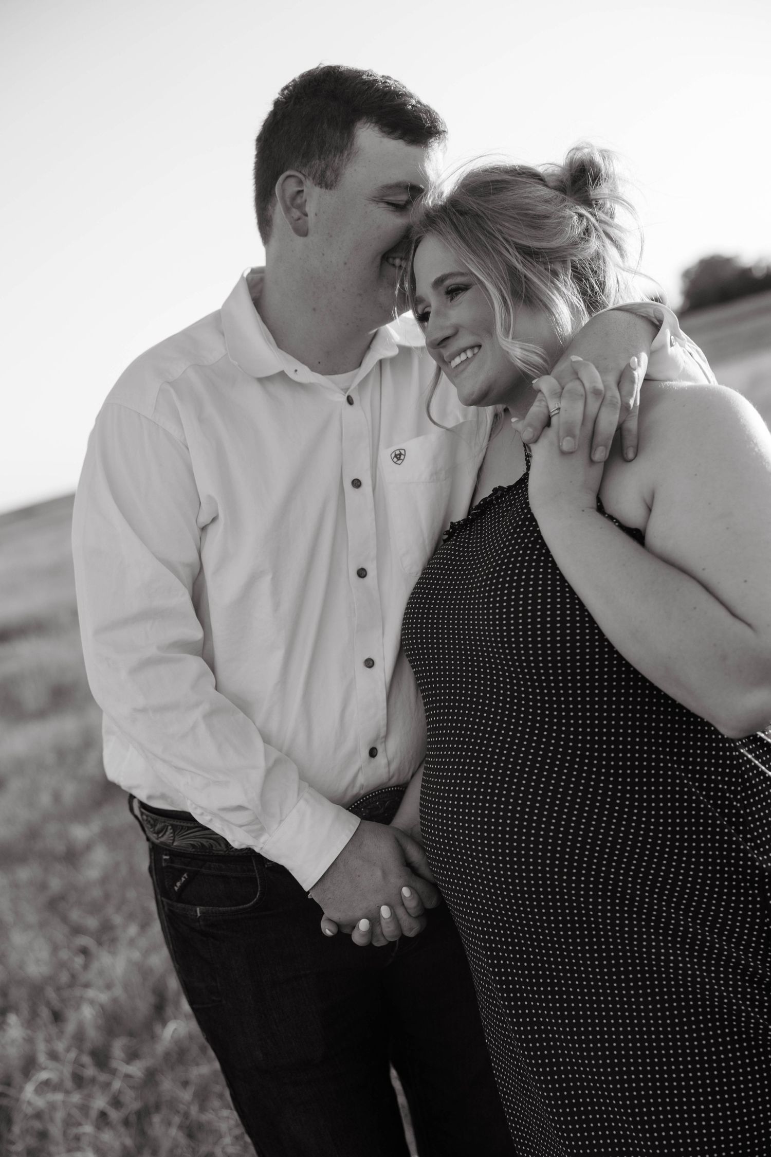 Black and white portrait of smiling pregnant woman and partner holding hands in open field at dusk.