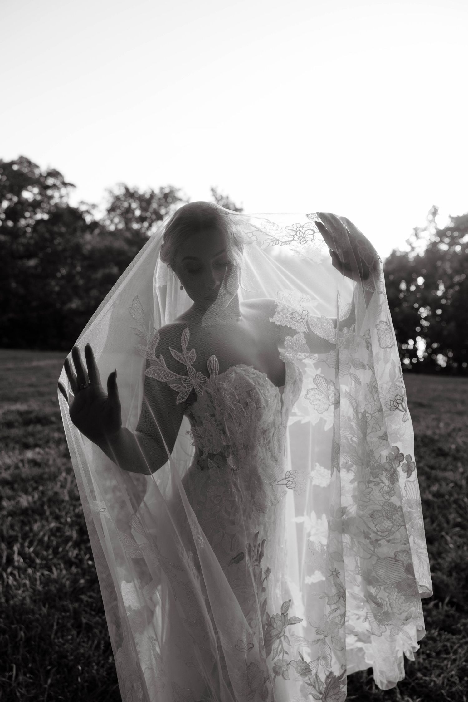 A black and white photo of a bride lifting her lace veil in a sunlit outdoor setting.