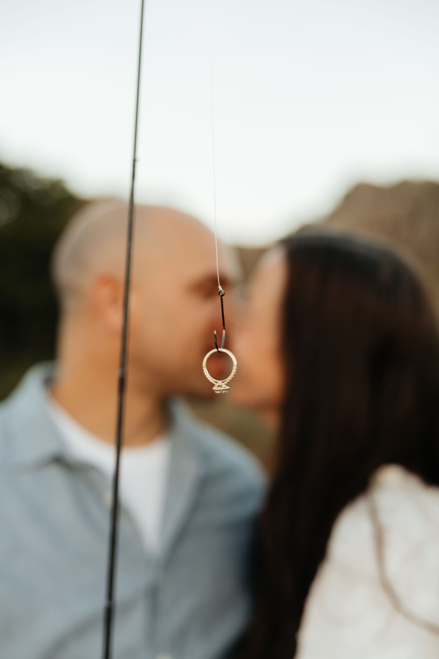 Engagement ring hanging from fishing line with couple kissing softly in background.