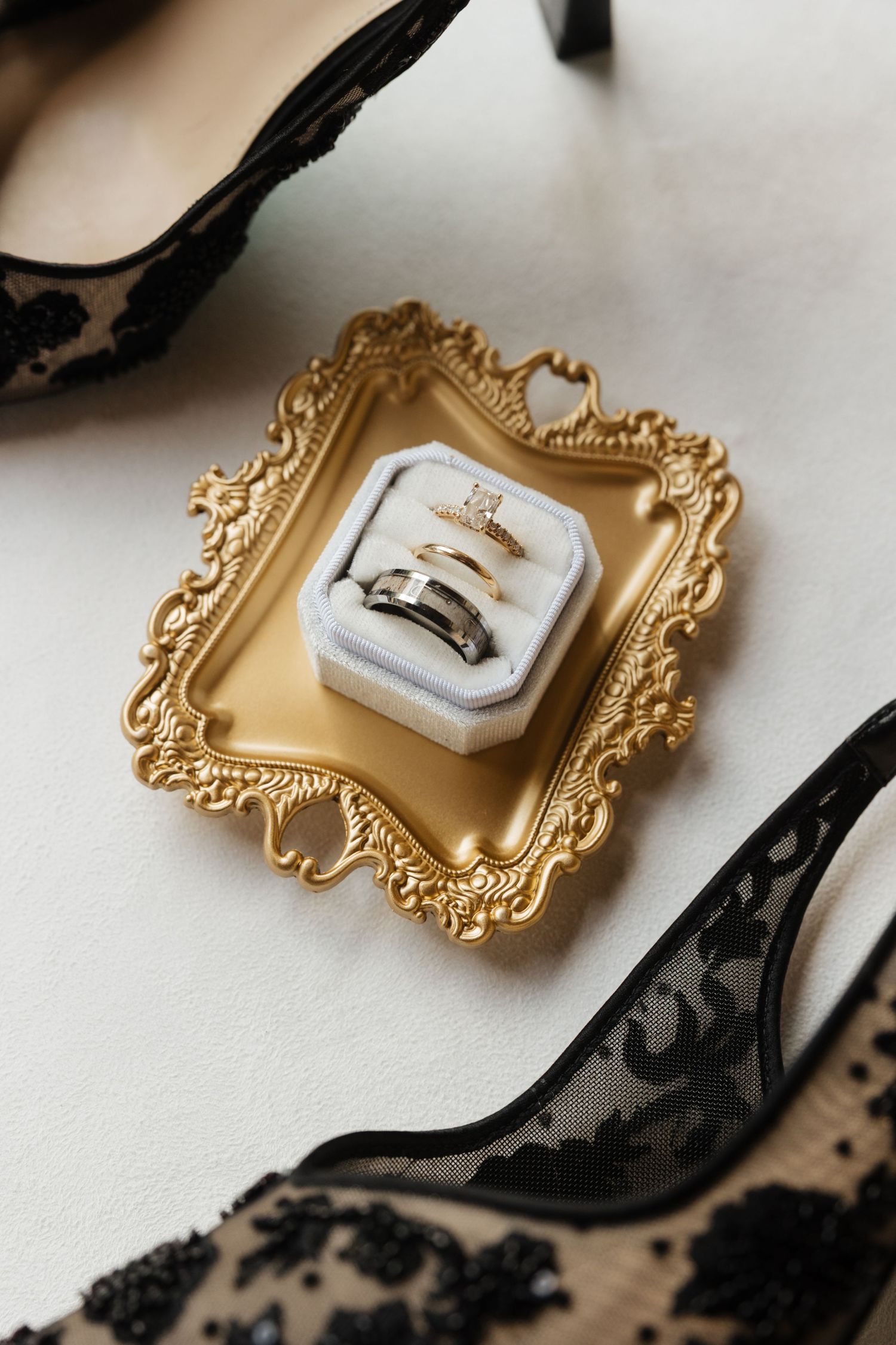Wedding rings and diamond engagement ring in white velvet box on gold ornate tray beside black heels.