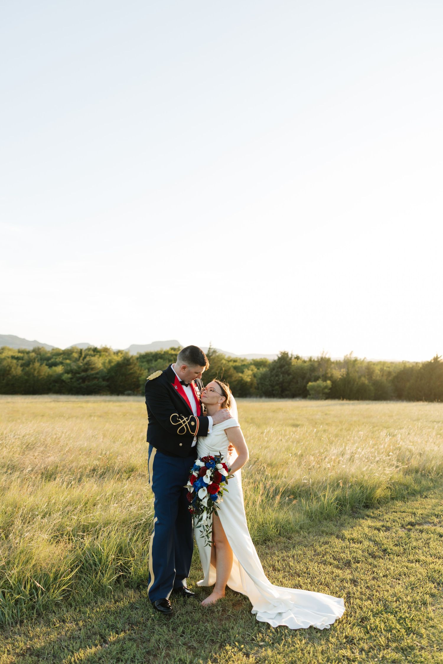 Bride and groom share a romantic moment in a golden field, she holds a red, white, and blue bouquet as he wears a military dress uniform.