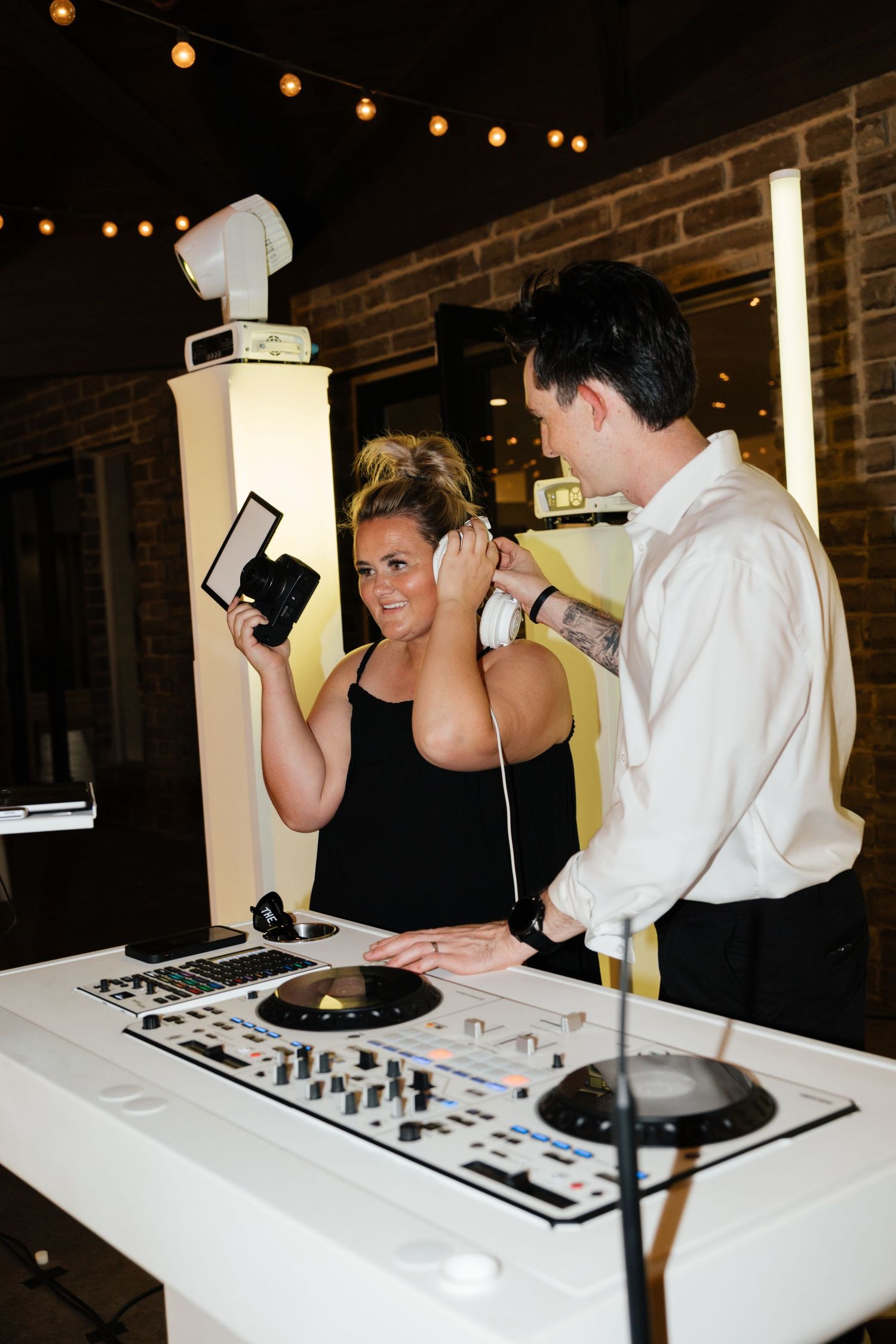 Bride tries on DJ headphones while holding a camera at the DJ booth during her wedding reception.