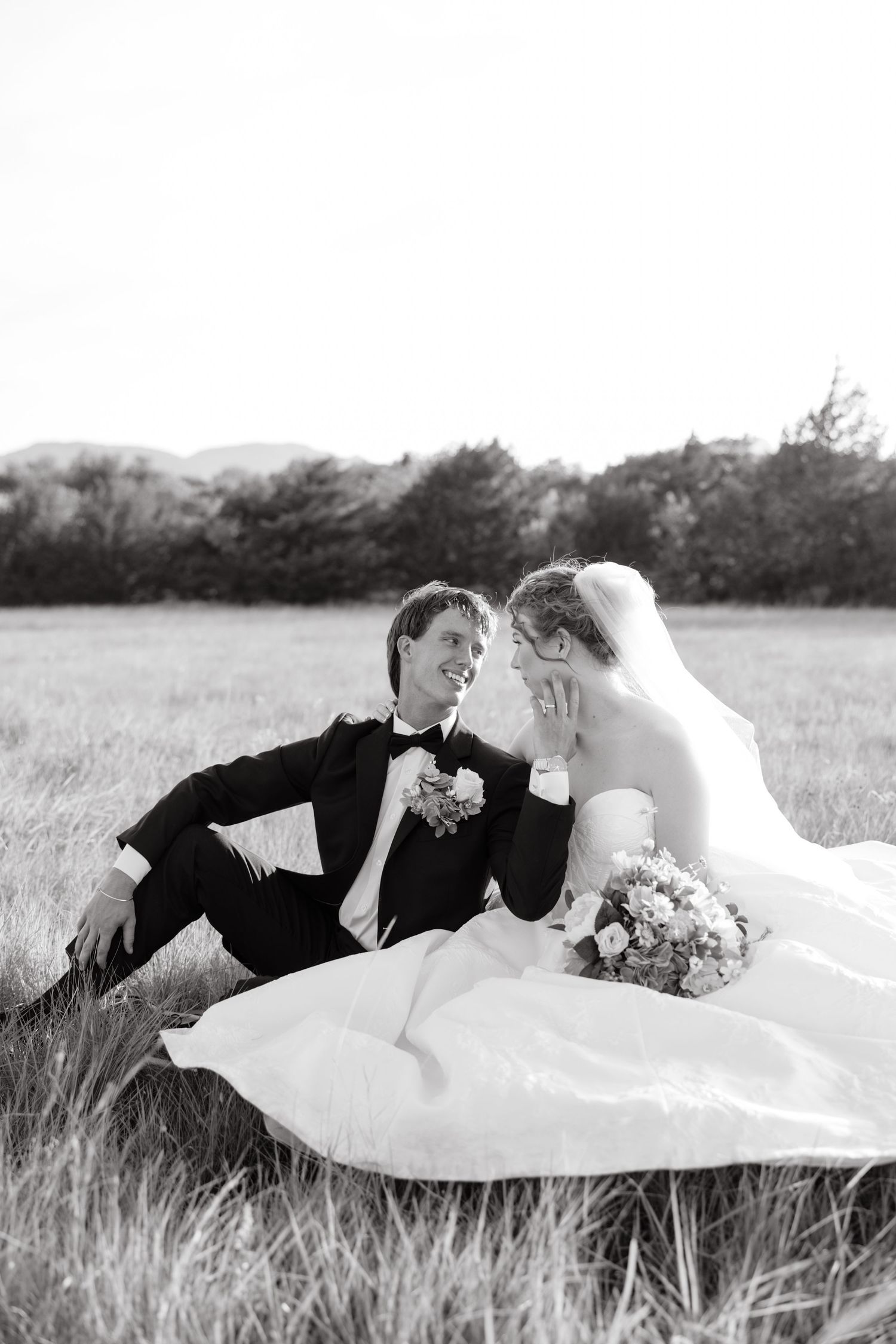 Black and white photo of bride and groom sitting together in a grassy field, smiling at each other on their wedding day.