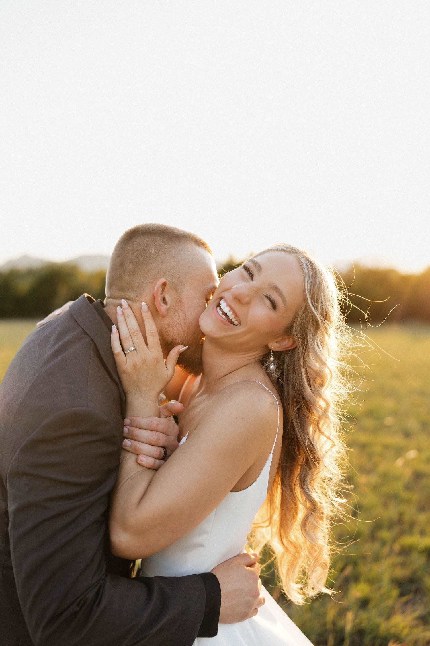 Joyful blonde bride in white dress laughing as groom kisses her cheek at golden hour in open field.