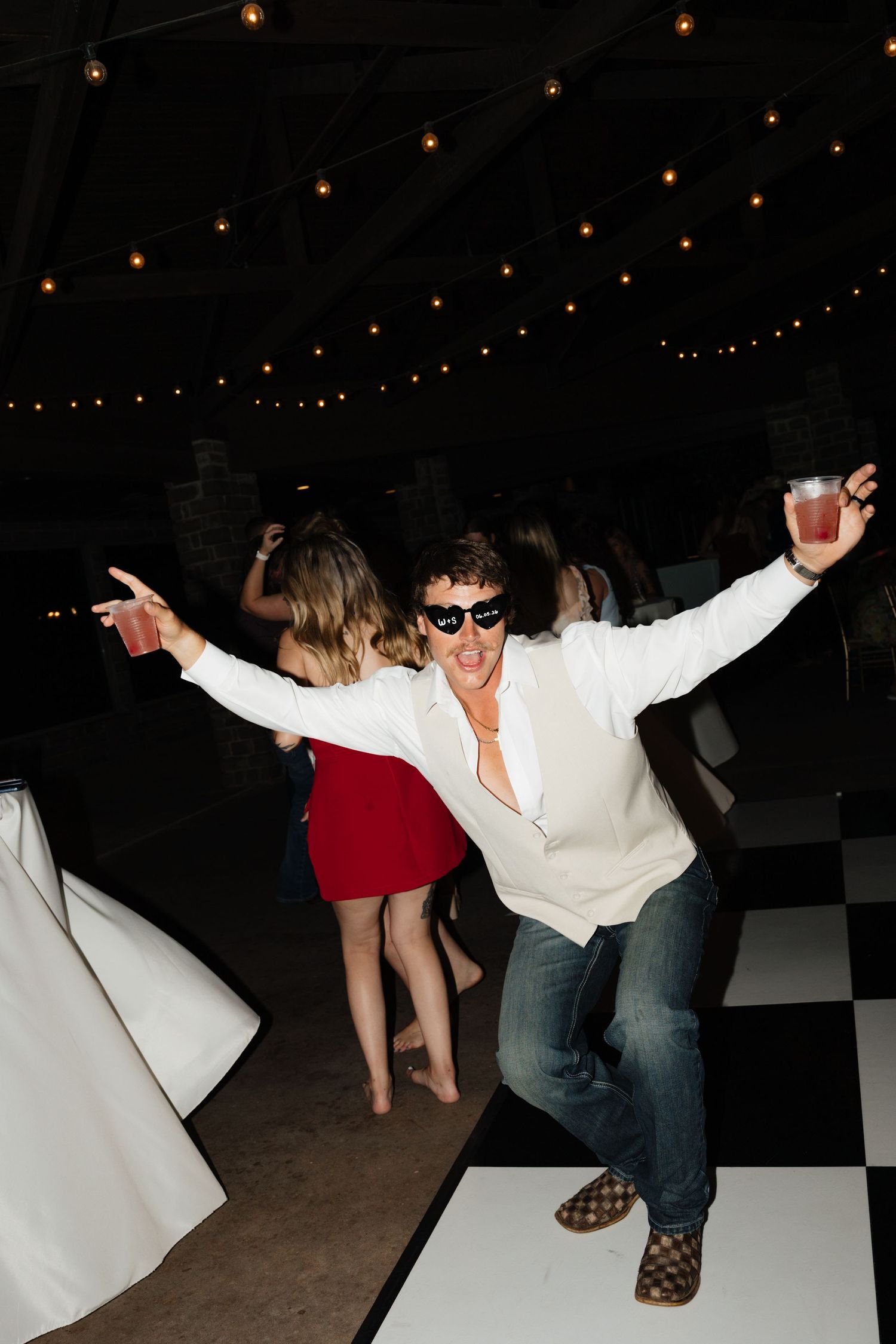 Energetic wedding guest in sunglasses dances with arms wide open on a checkered dance floor at the reception.