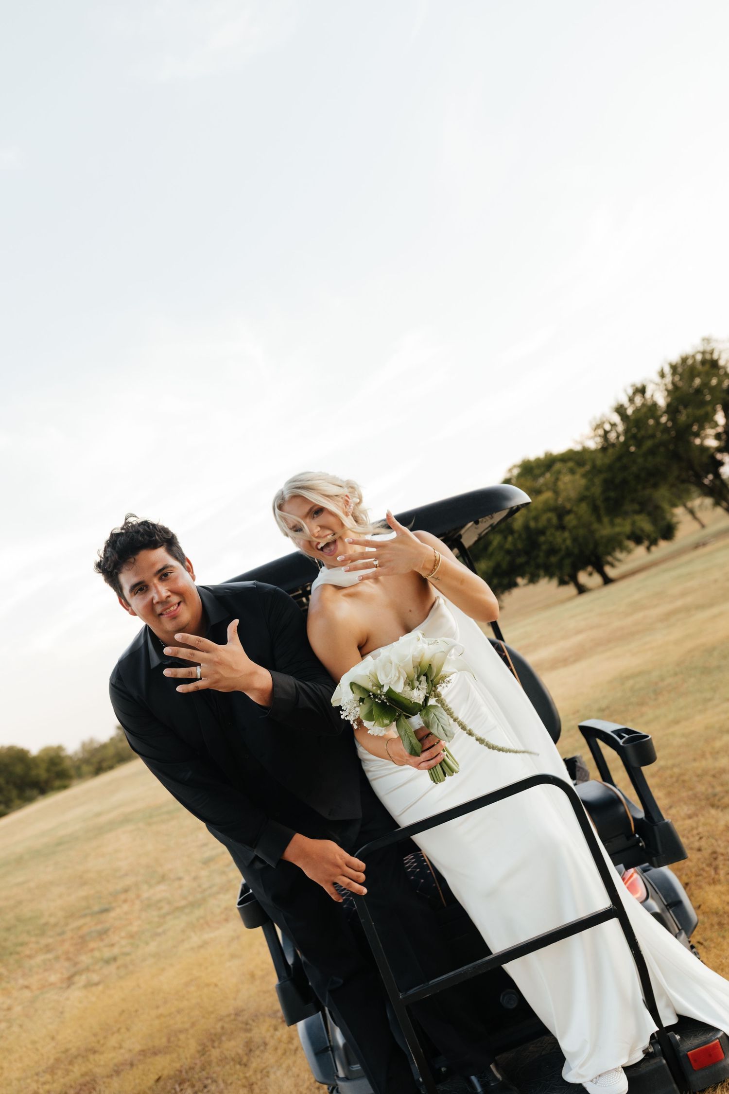 Smiling bride and groom pose playfully beside a golf cart in an open field on their wedding day.