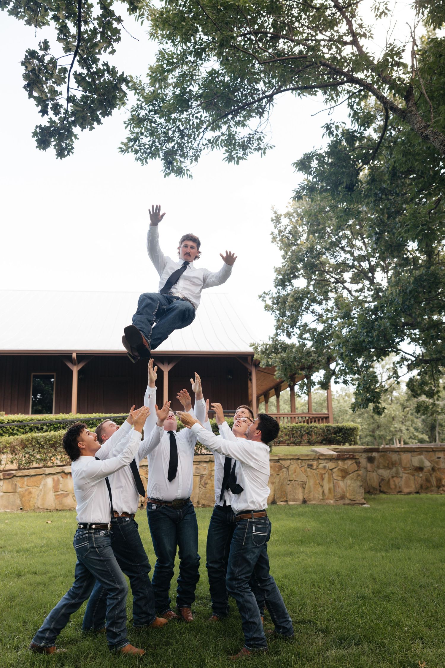 Groomsmen tossing groom into the air in celebration outside rustic wedding venue surrounded by trees.