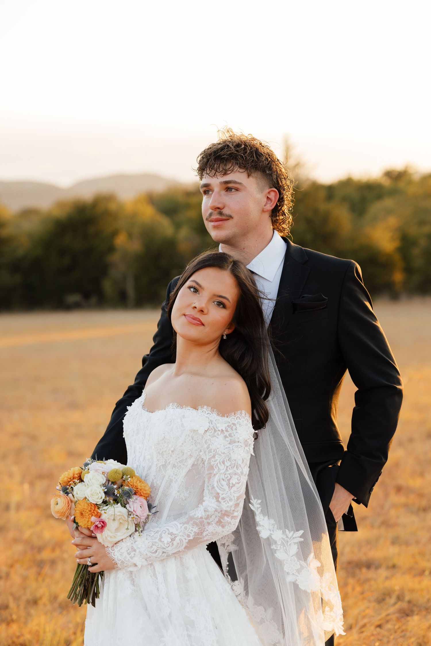A bride and groom pose together in a golden field at sunset during their wedding.