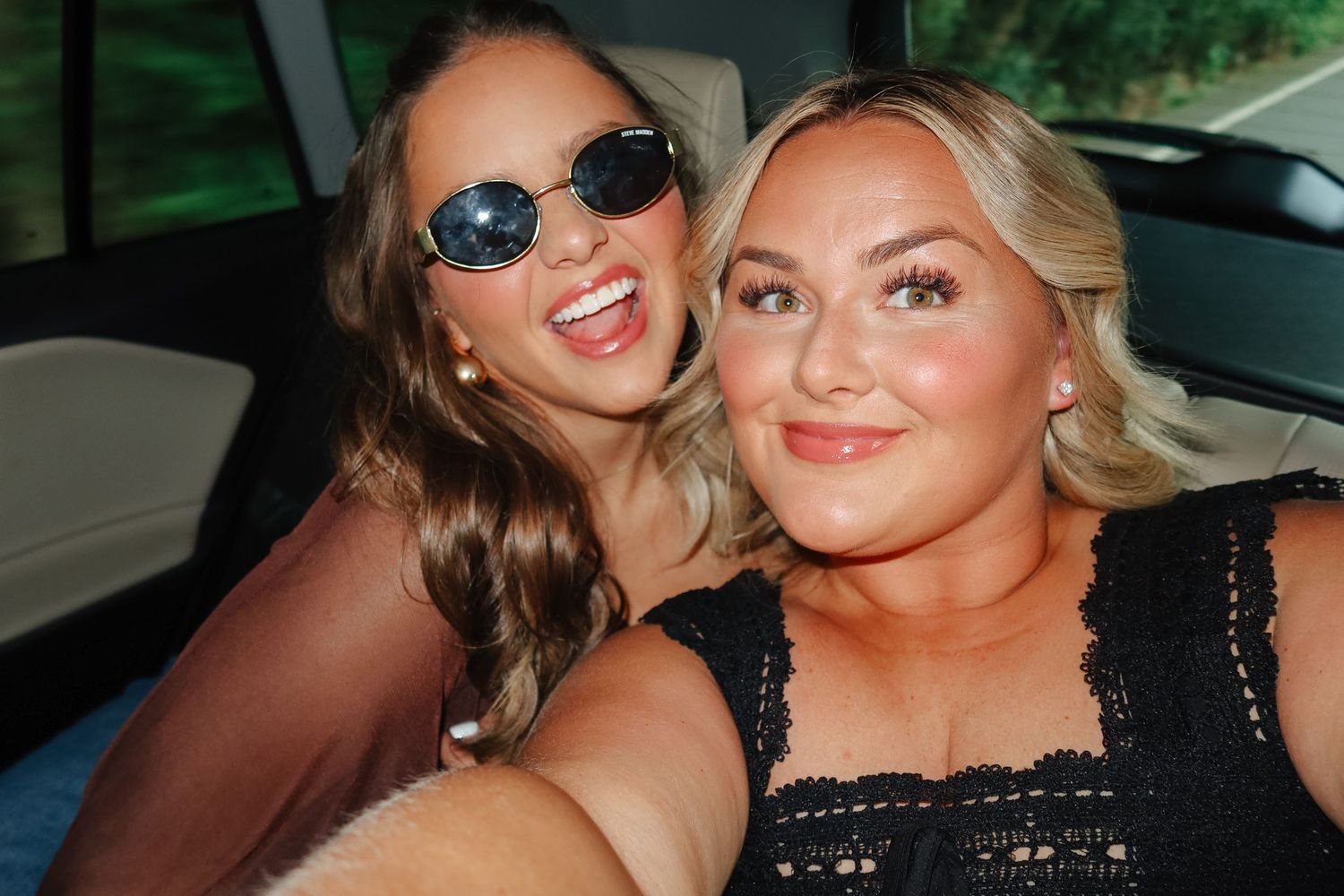 Two smiling women take a fun selfie together inside a car, one wearing sunglasses.