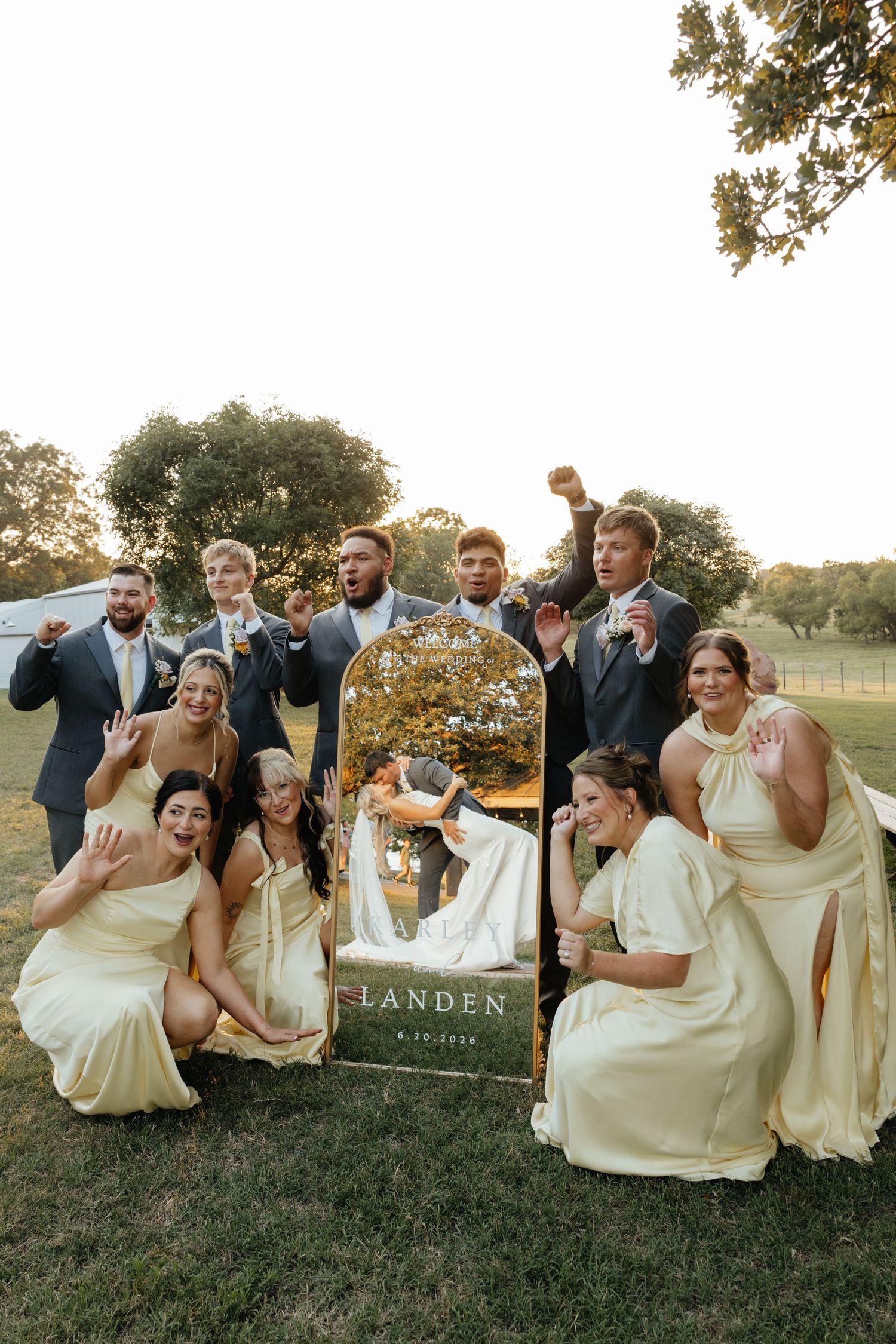 Wedding party posing playfully around decorative mirror sign at outdoor golden hour reception.