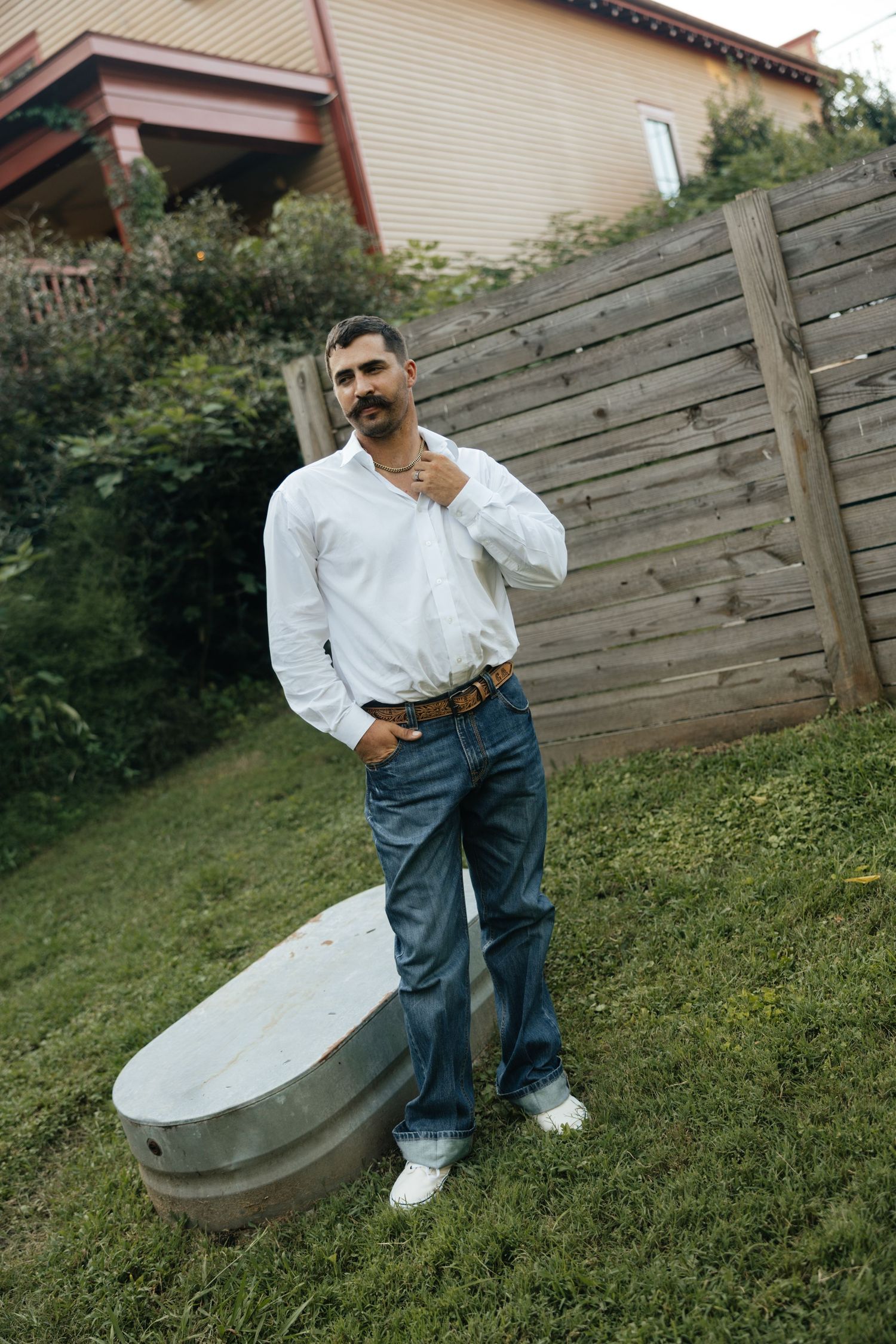 Man in white shirt and jeans posing outdoors near wooden fence and metal trough in backyard.