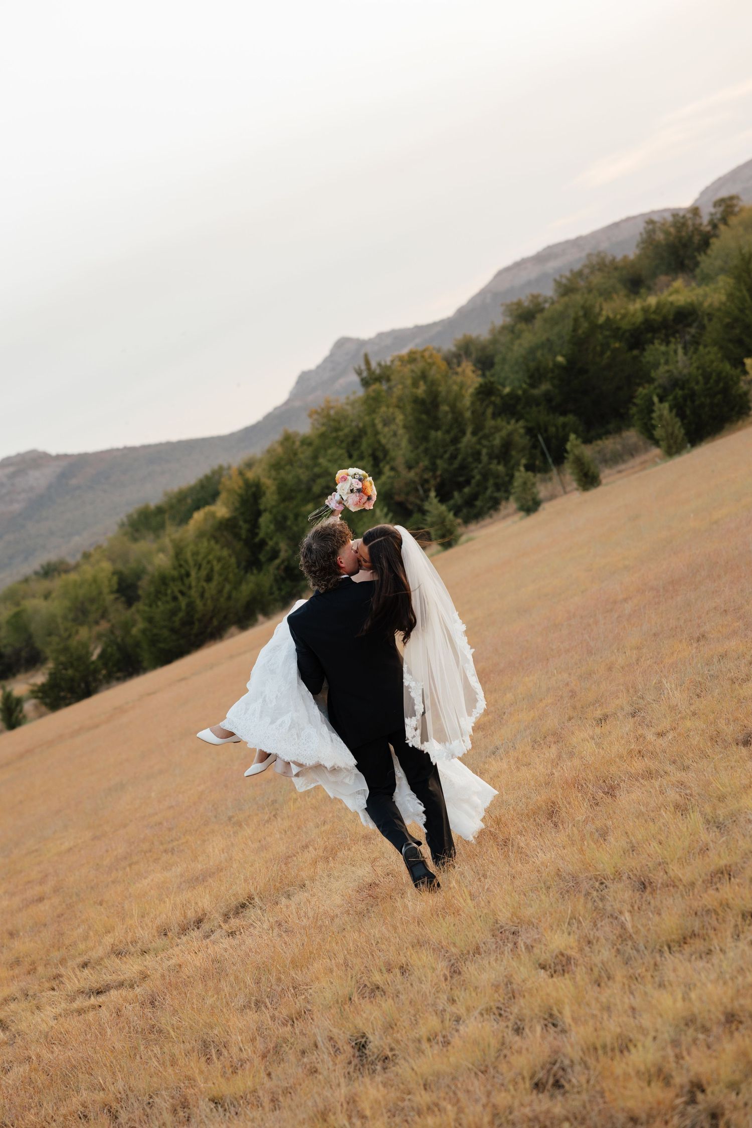 Groom carrying bride through golden mountain meadow, her veil and bouquet flowing in wind.